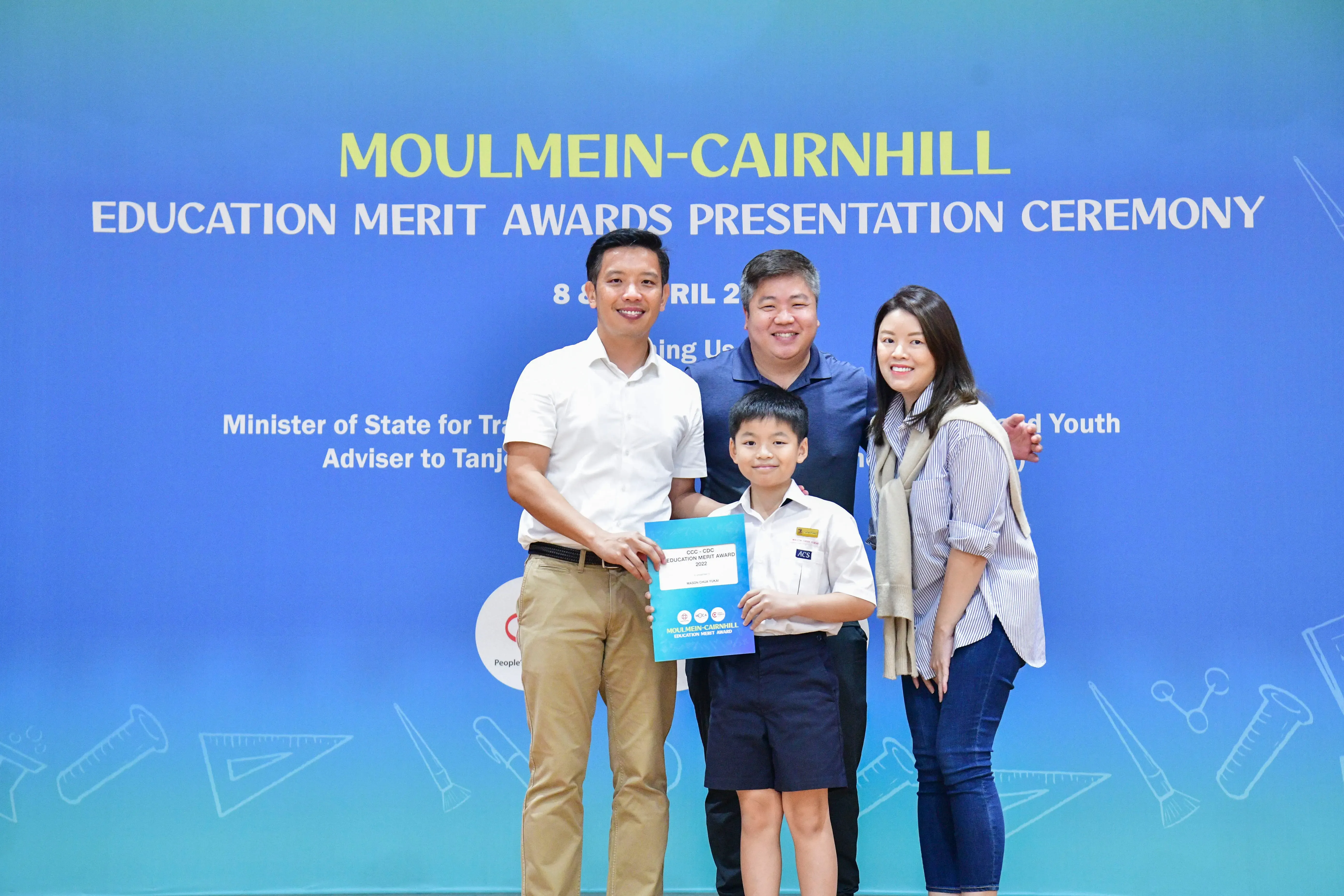 Four people pose before a Moulmein-Cairnhill Education Merit Awards banner, with a boy holding an award certificate.