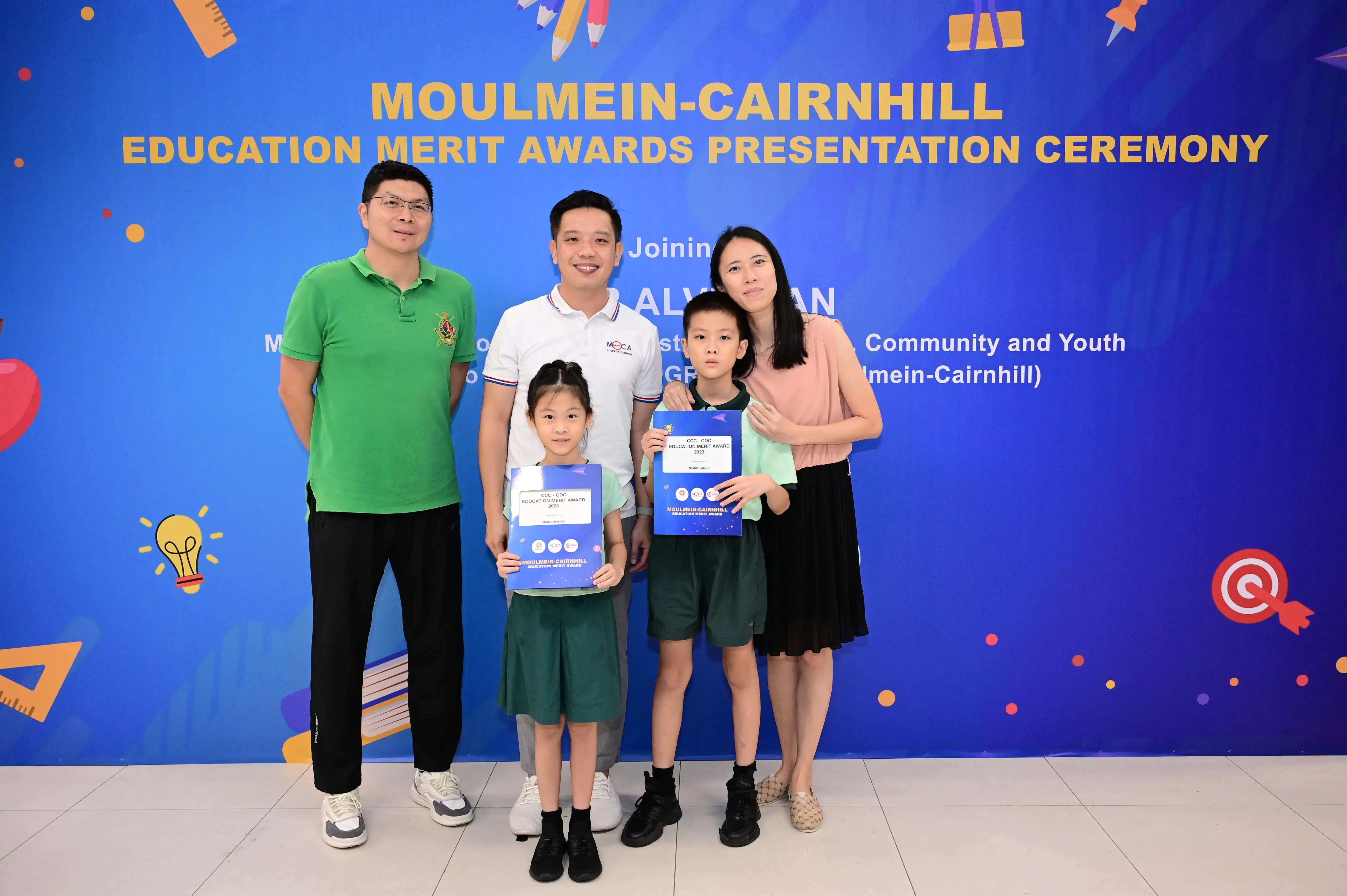 Five people pose before a Moulmein-Cairnhill Education Merit Awards banner, two children holding award certificates.
