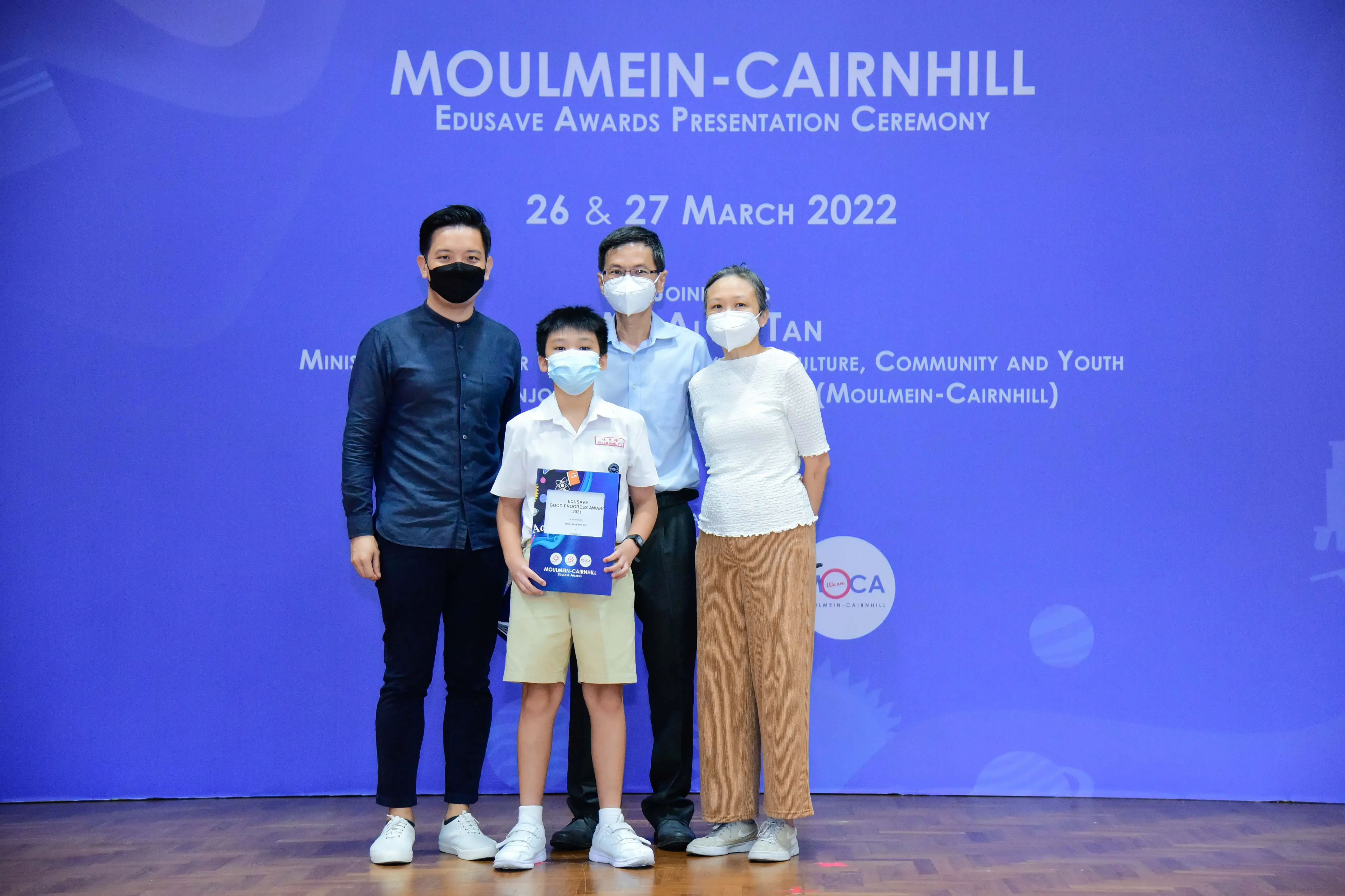 Four masked people pose at the Moulmein-Cairnhill Edusave Awards Ceremony on 26-27 March 2022, with a boy holding a certificate.
