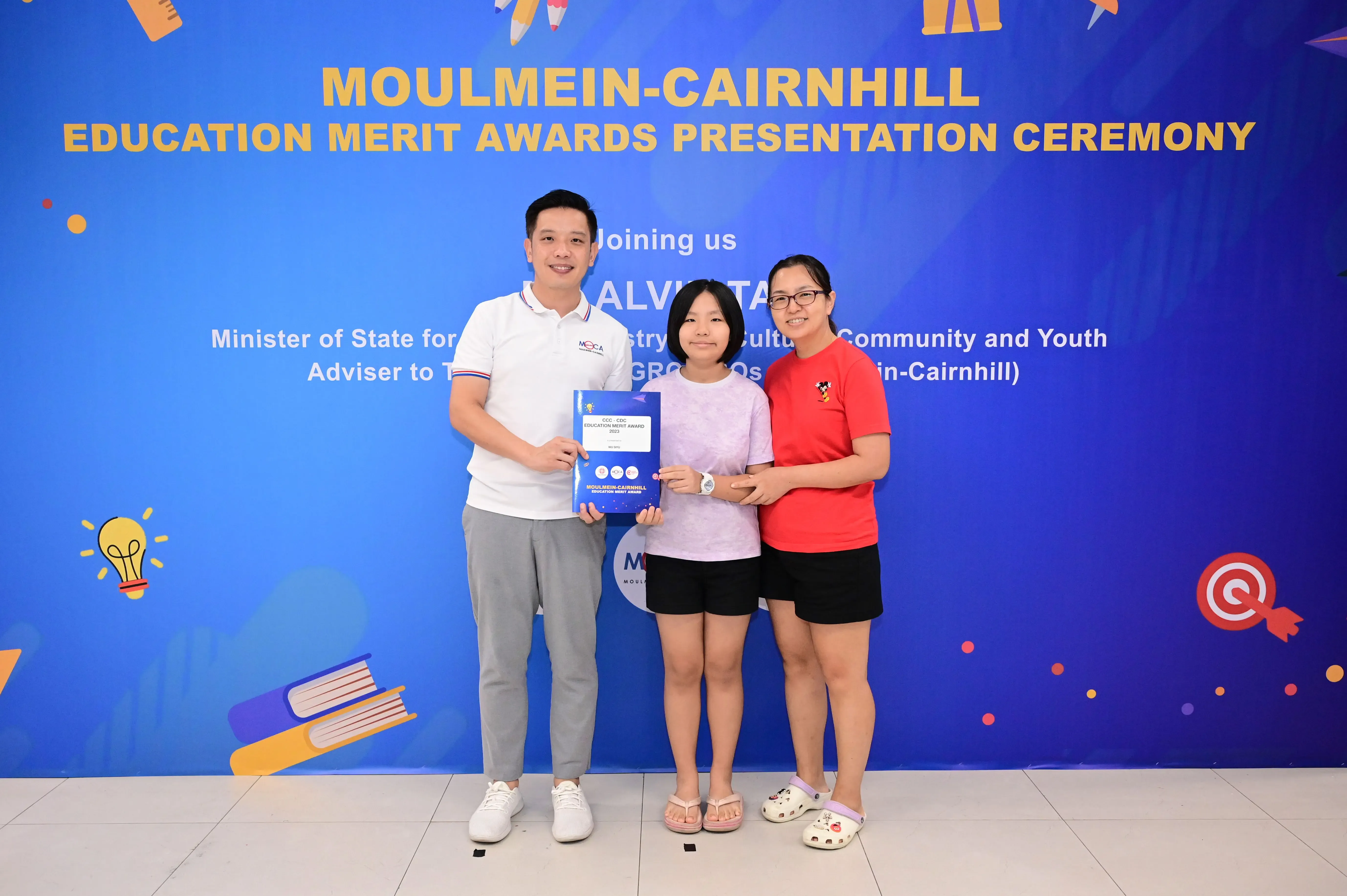 Three people pose holding an award booklet in front of a Moulmein-Cairnhill Education Merit Awards banner.
