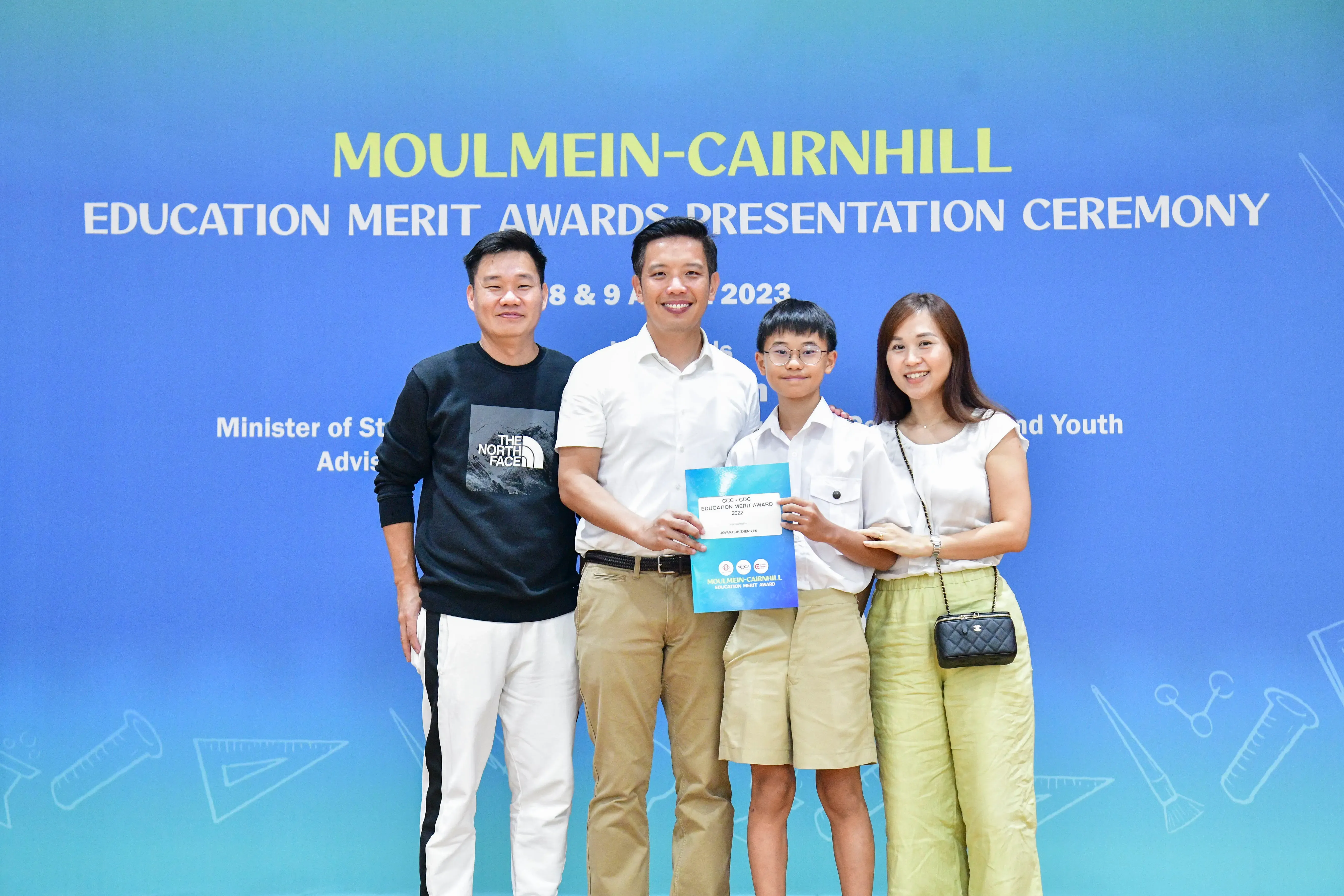 Four people smile in front of a blue Moulmein-Cairnhill Education Merit Awards Presentation Ceremony banner, holding a certificate.