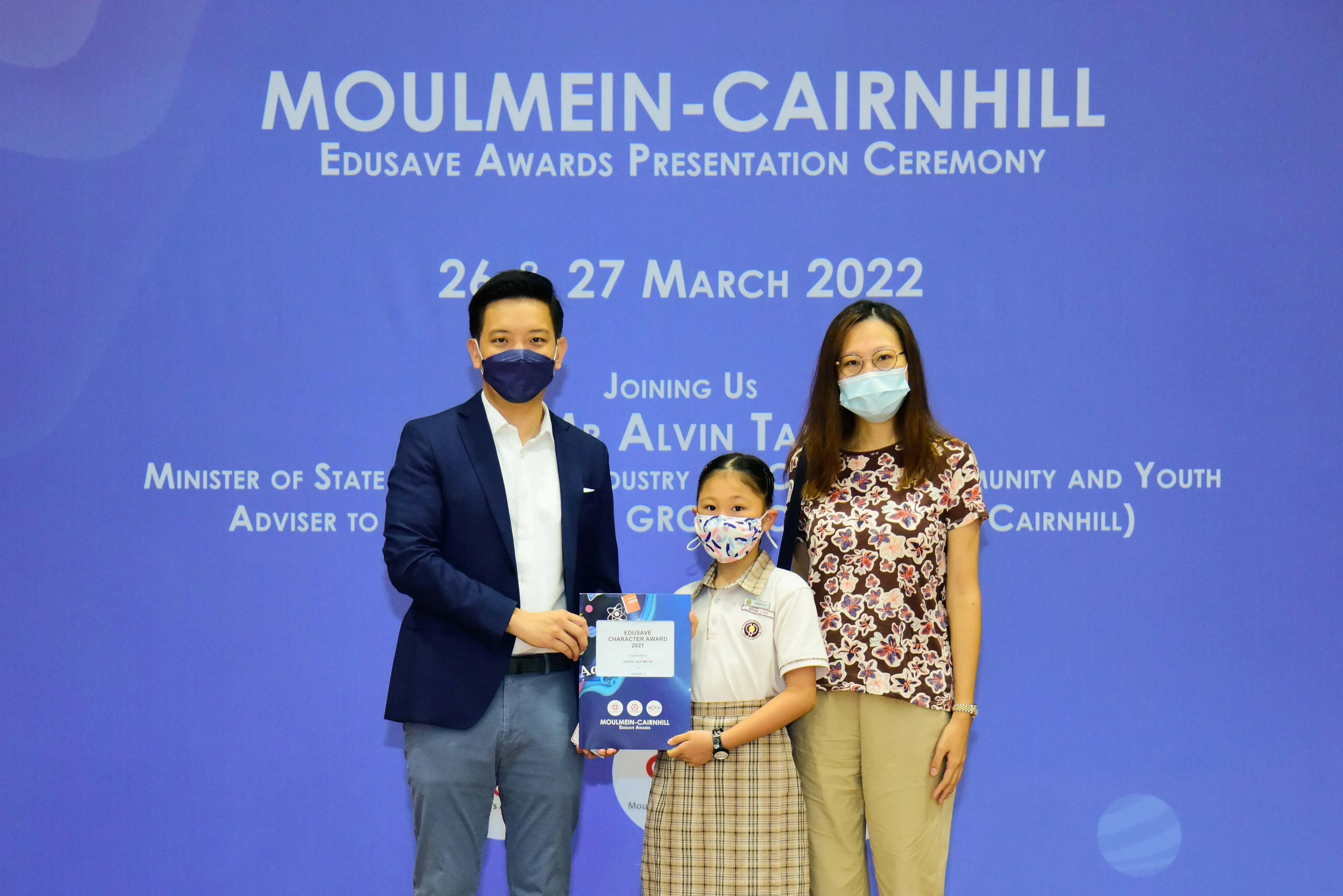 Two masked adults and a masked child holding an award certificate pose at the Moulmein-Cairnhill Edusave Awards Ceremony 2022.
