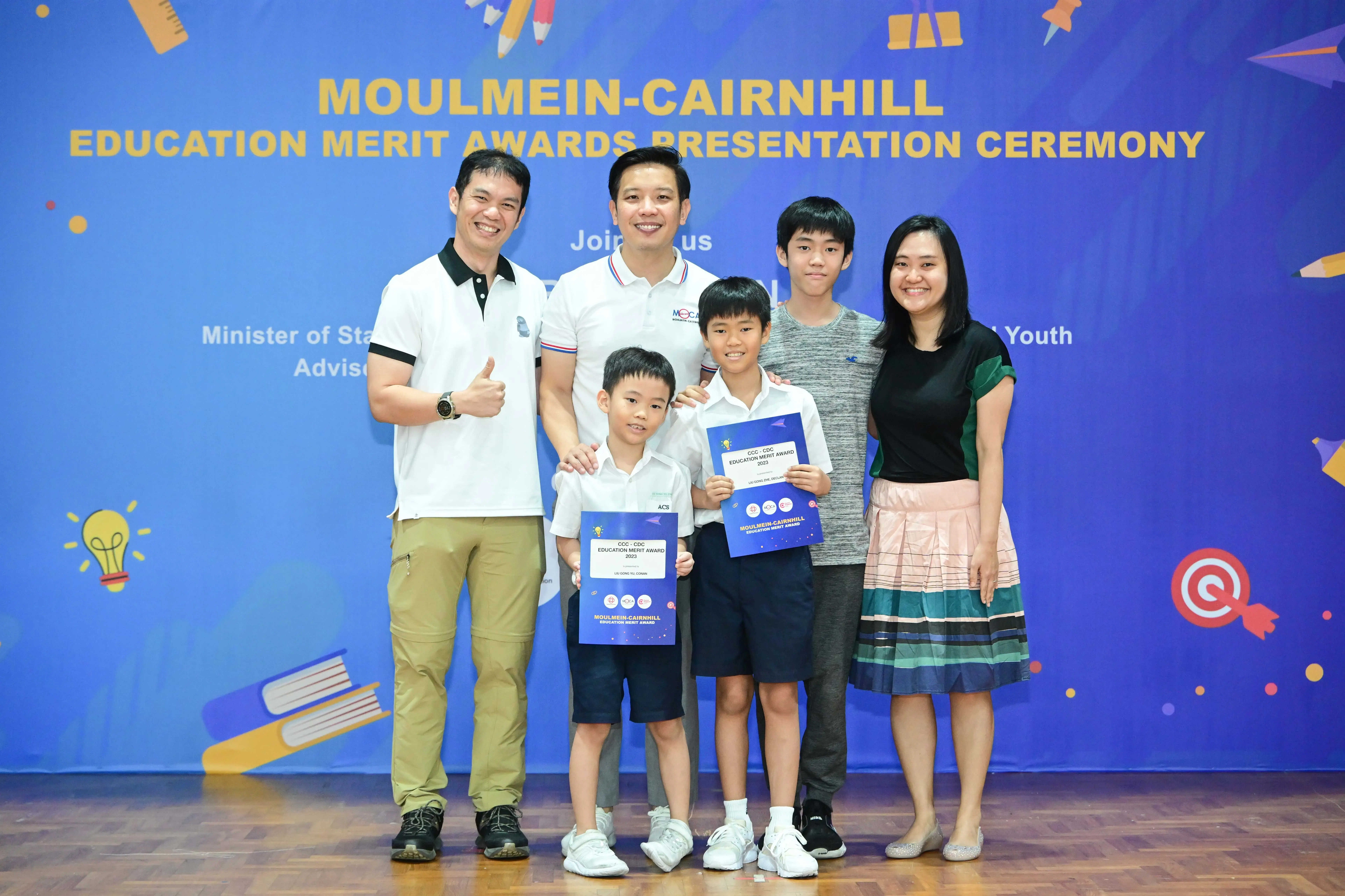 Five people pose at the Moulmein-Cairnhill Education Merit Awards ceremony, two children holding award certificates.