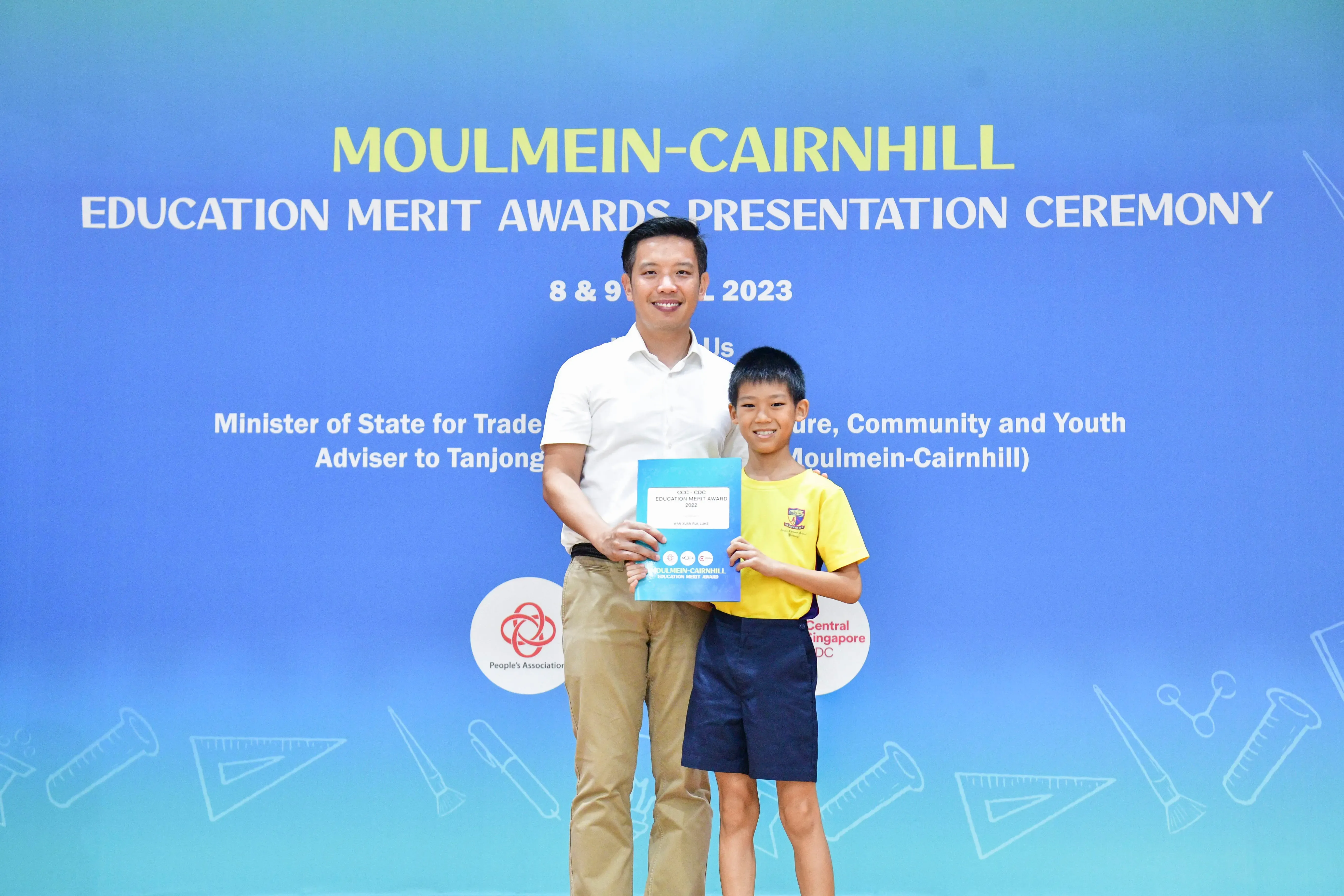 An adult and a young boy hold a certificate in front of a Moulmein-Cairnhill Education Merit Awards 2023 banner.