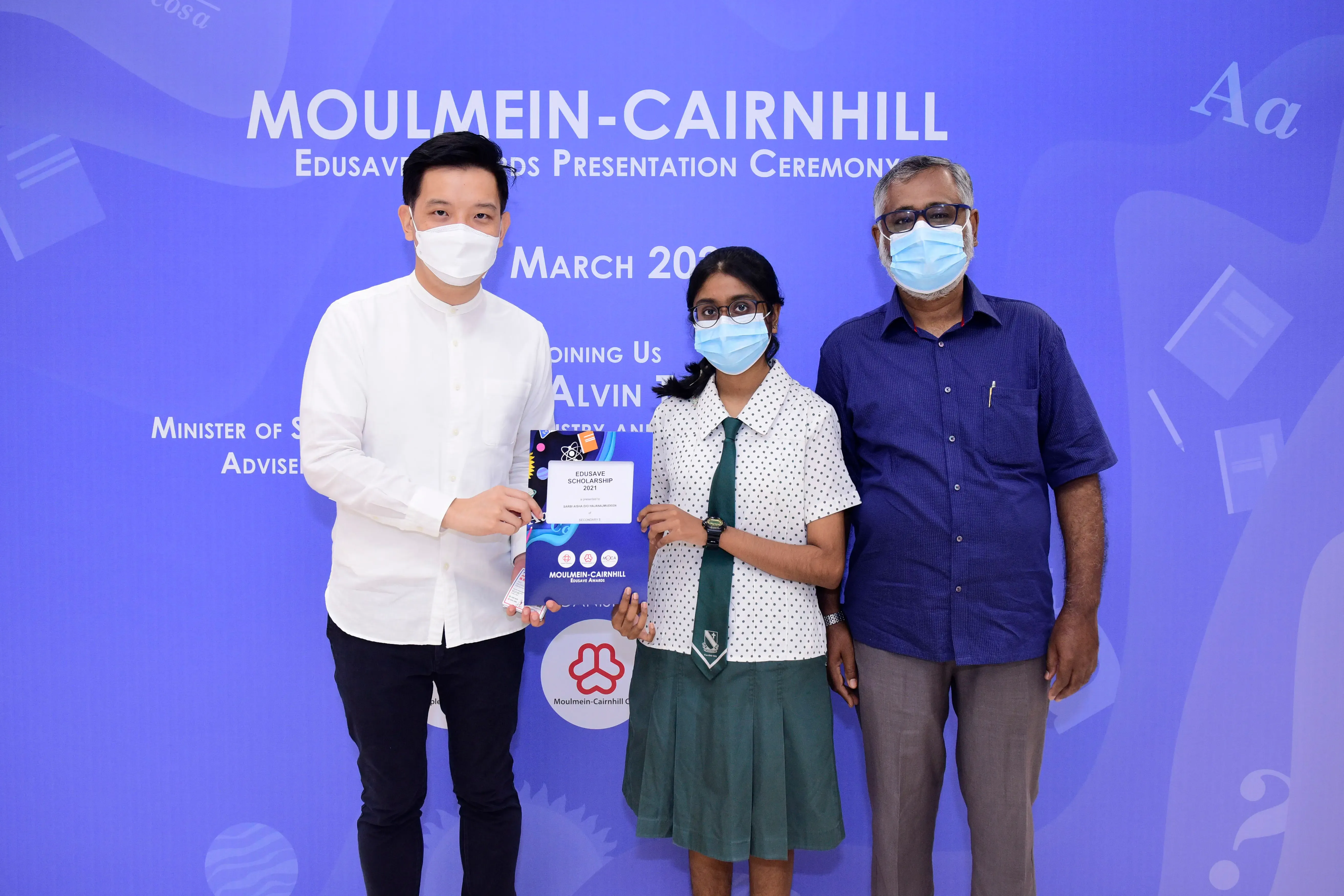 Three masked people pose at the Moulmein-Cairnhill Edusave Awards Presentation Ceremony, with a student holding a certificate.