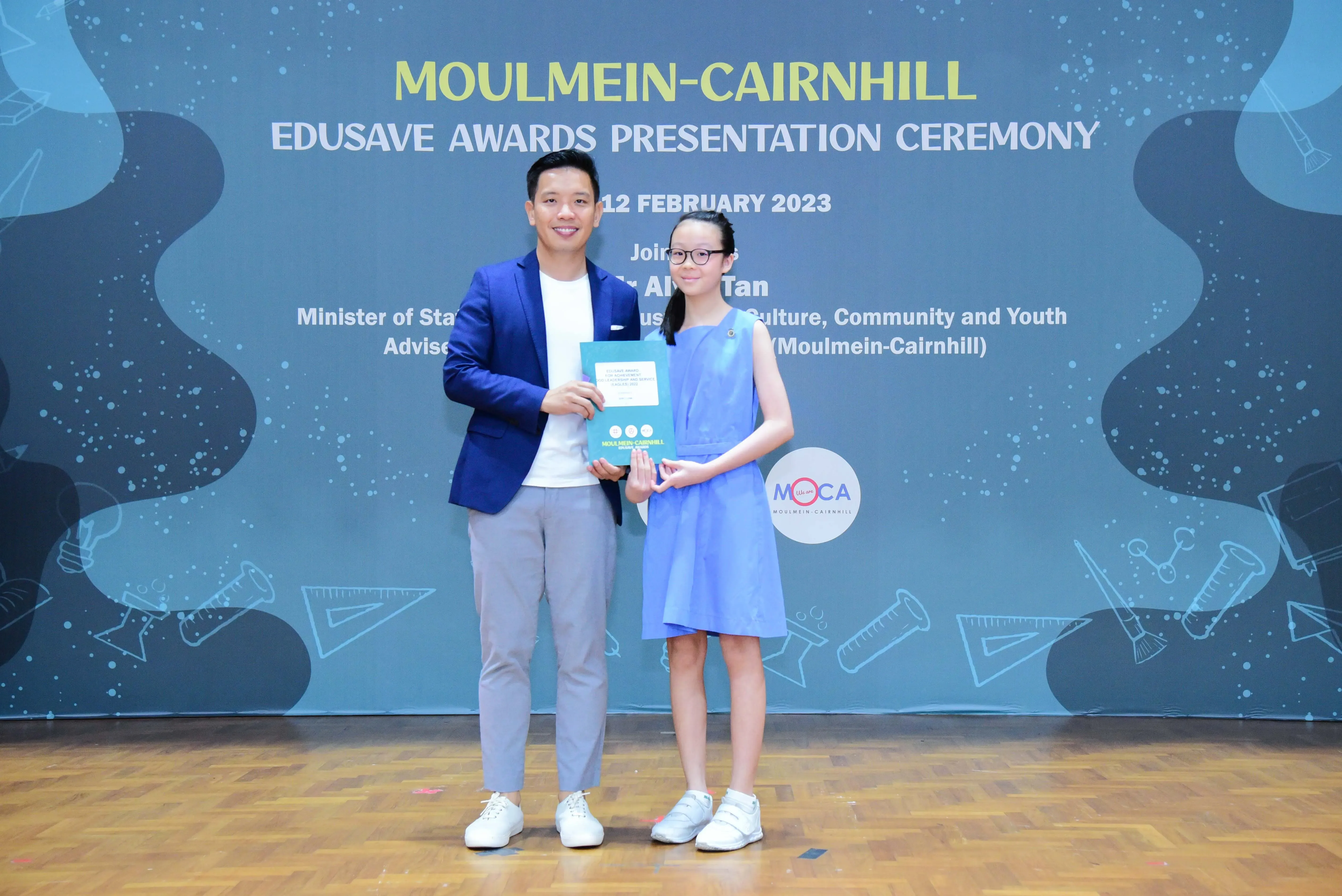 A man in a blue blazer and a young girl in a blue dress hold an award at the Moulmein-Cairnhill Edusave Awards Ceremony 2023.