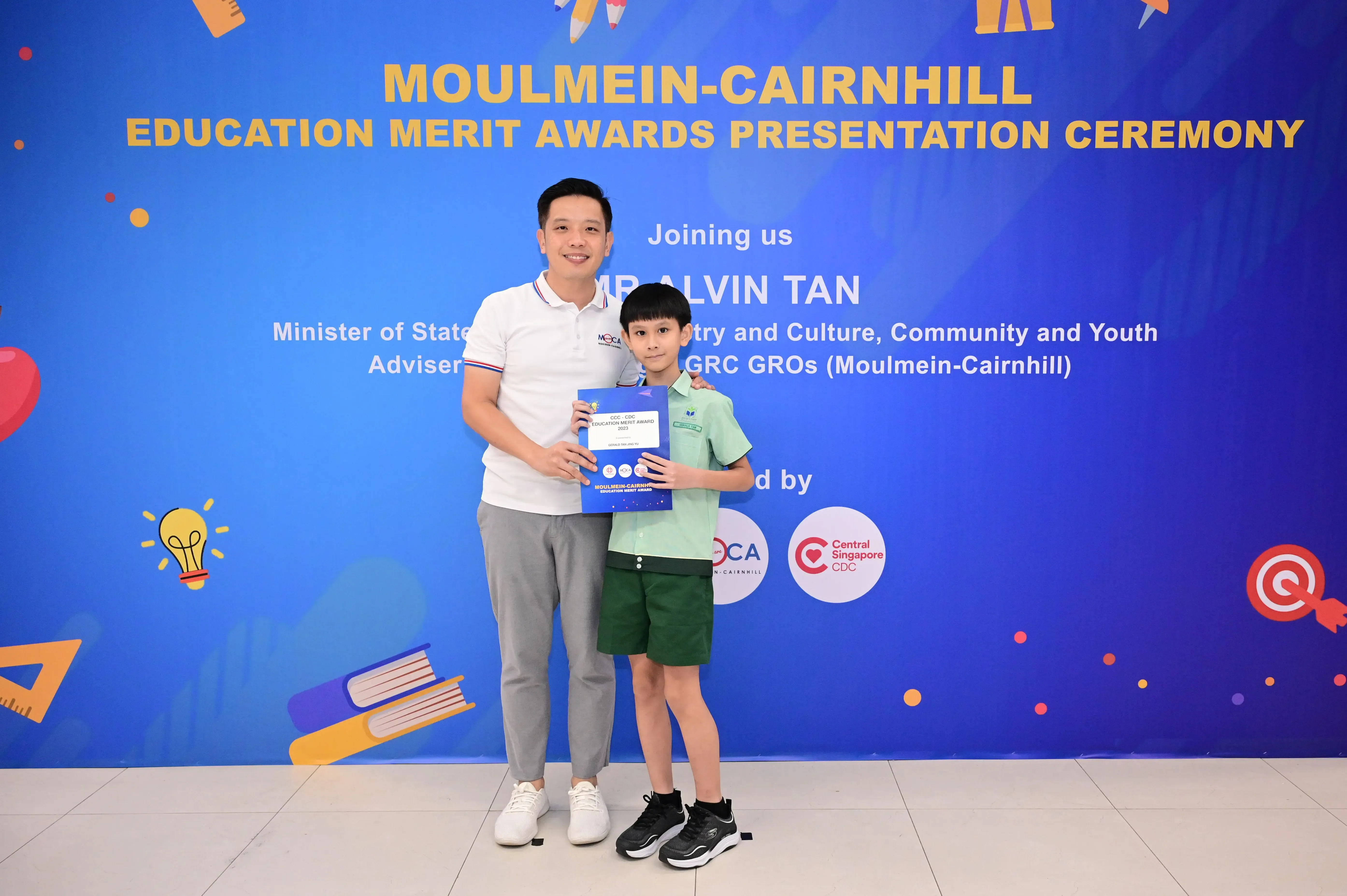 A man and a young boy hold a certificate in front of a Moulmein-Cairnhill Education Merit Awards banner.