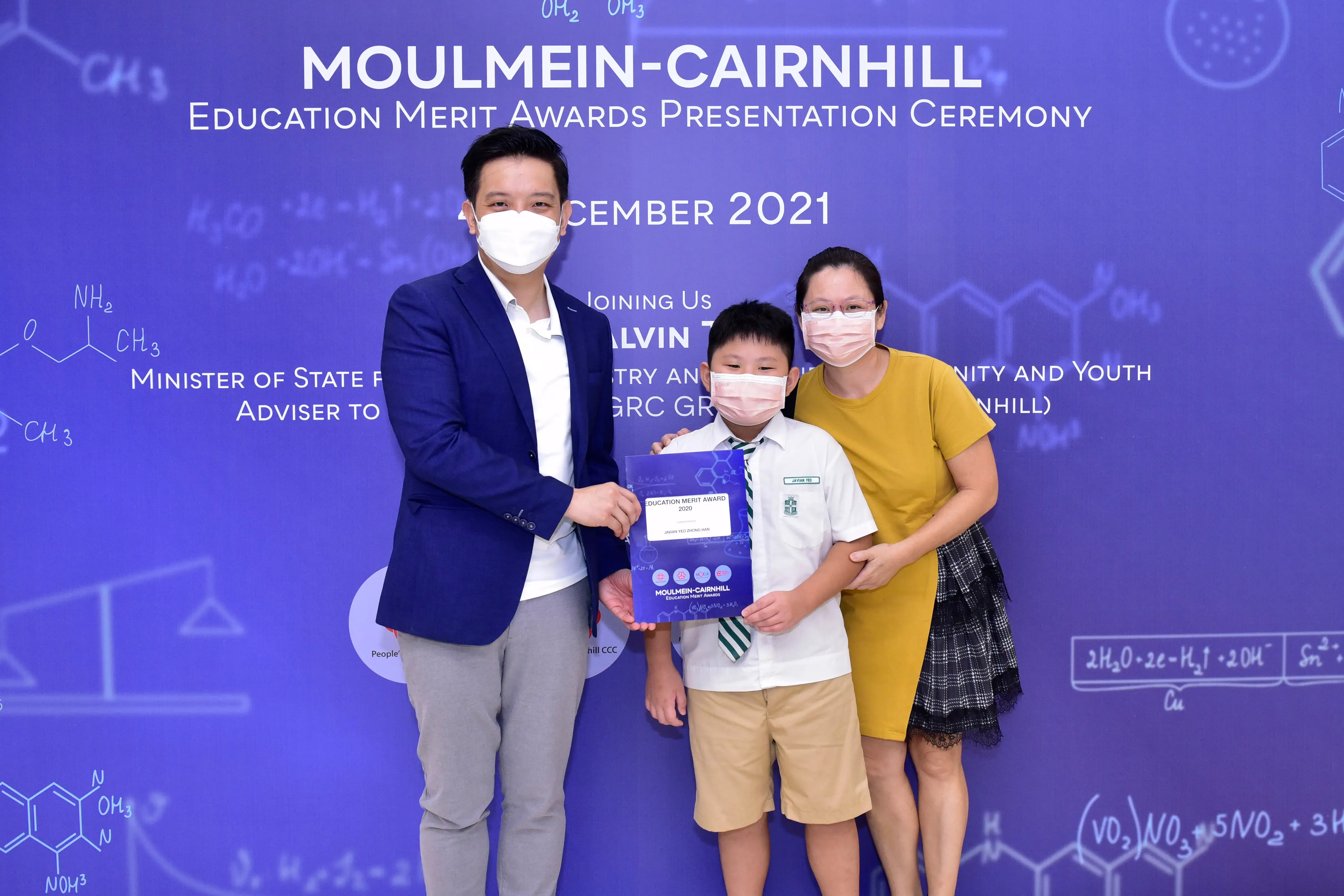 Three masked people pose at the Moulmein-Cairnhill Education Merit Awards Ceremony, with a boy holding an award certificate.