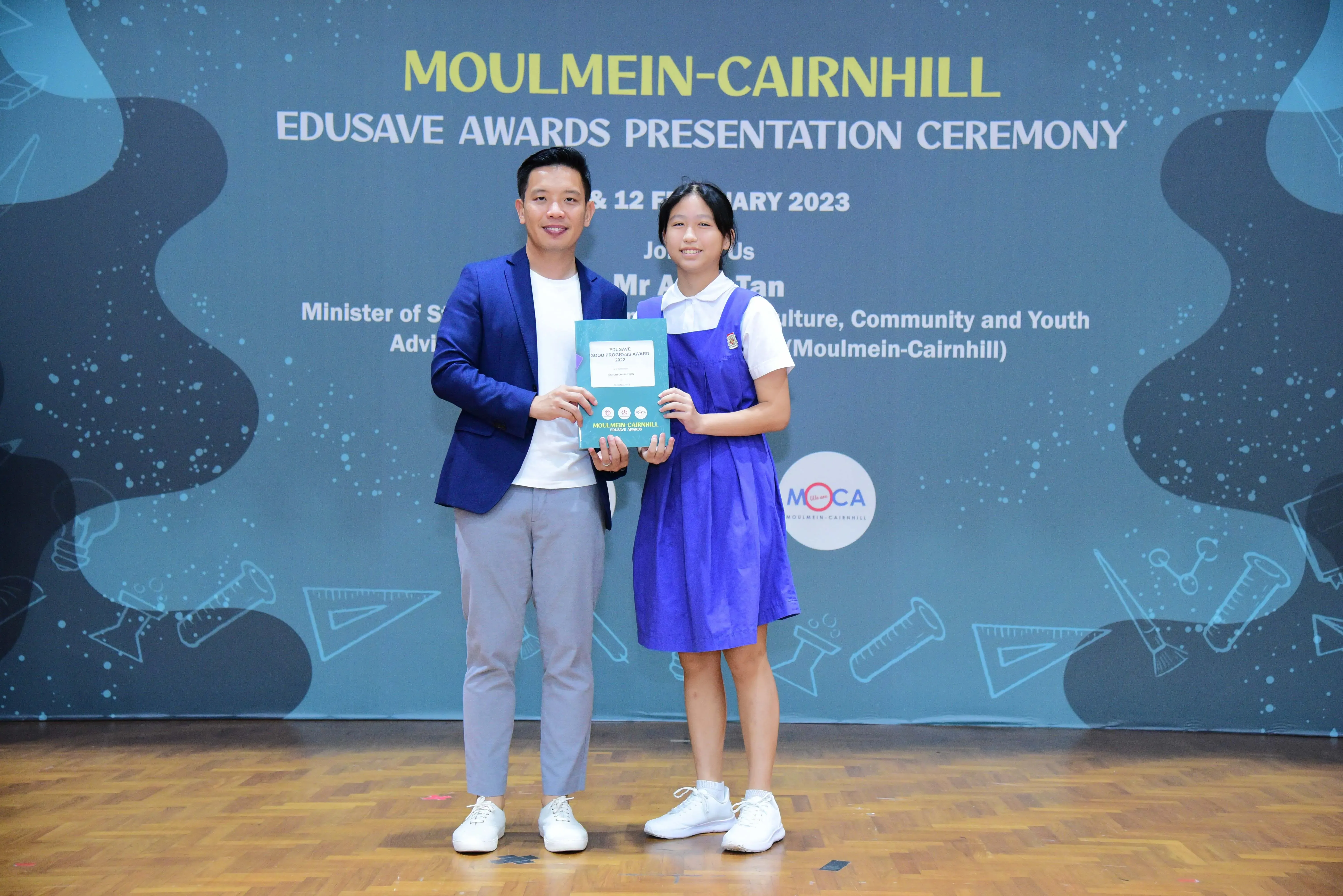 A man in a blue blazer and a girl in a school uniform hold an award at the Moulmein-Cairnhill Edusave Awards Ceremony 2023.