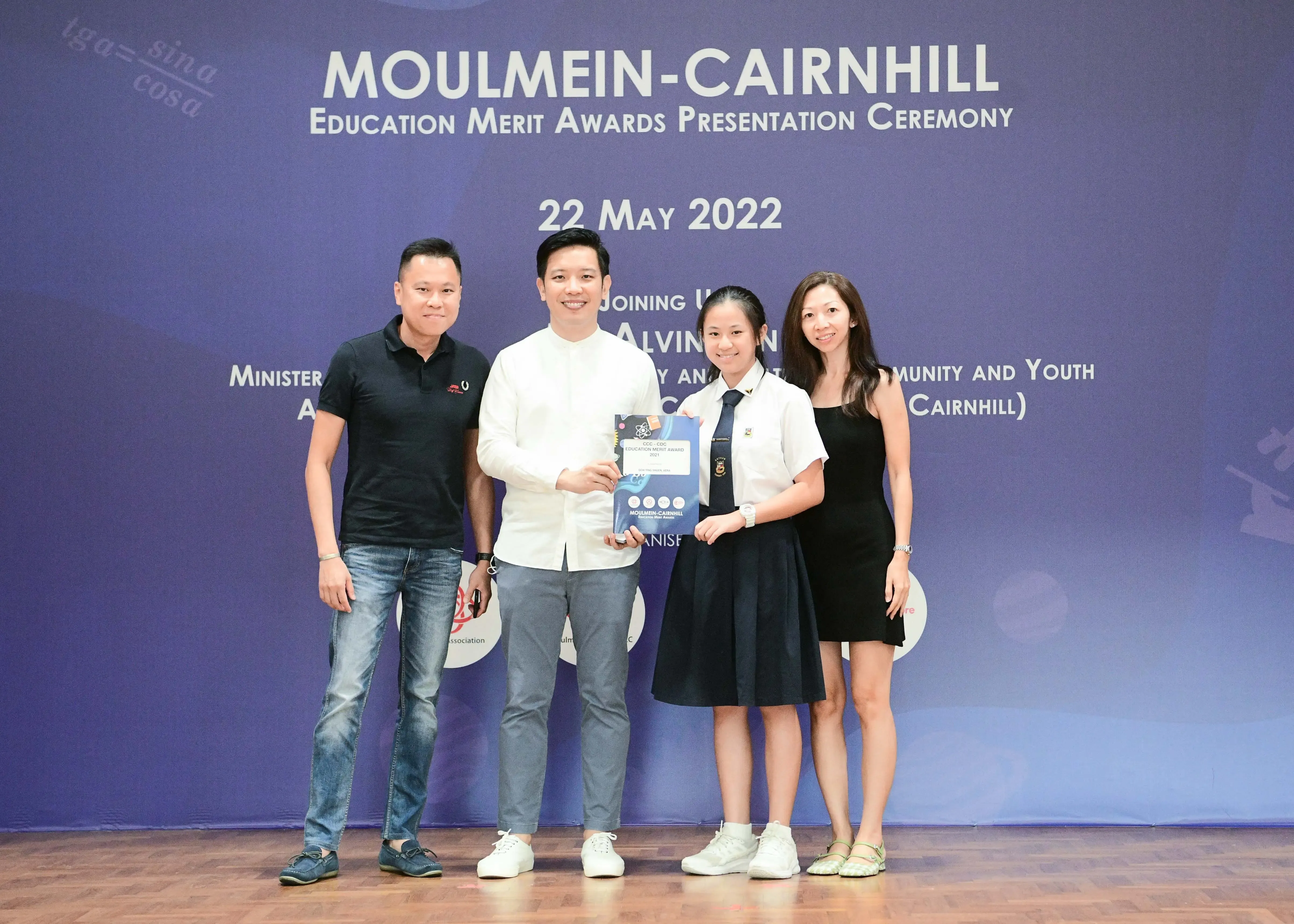 Four people pose in front of a Moulmein-Cairnhill Education Merit Awards Presentation Ceremony banner dated 22 May 2022.
