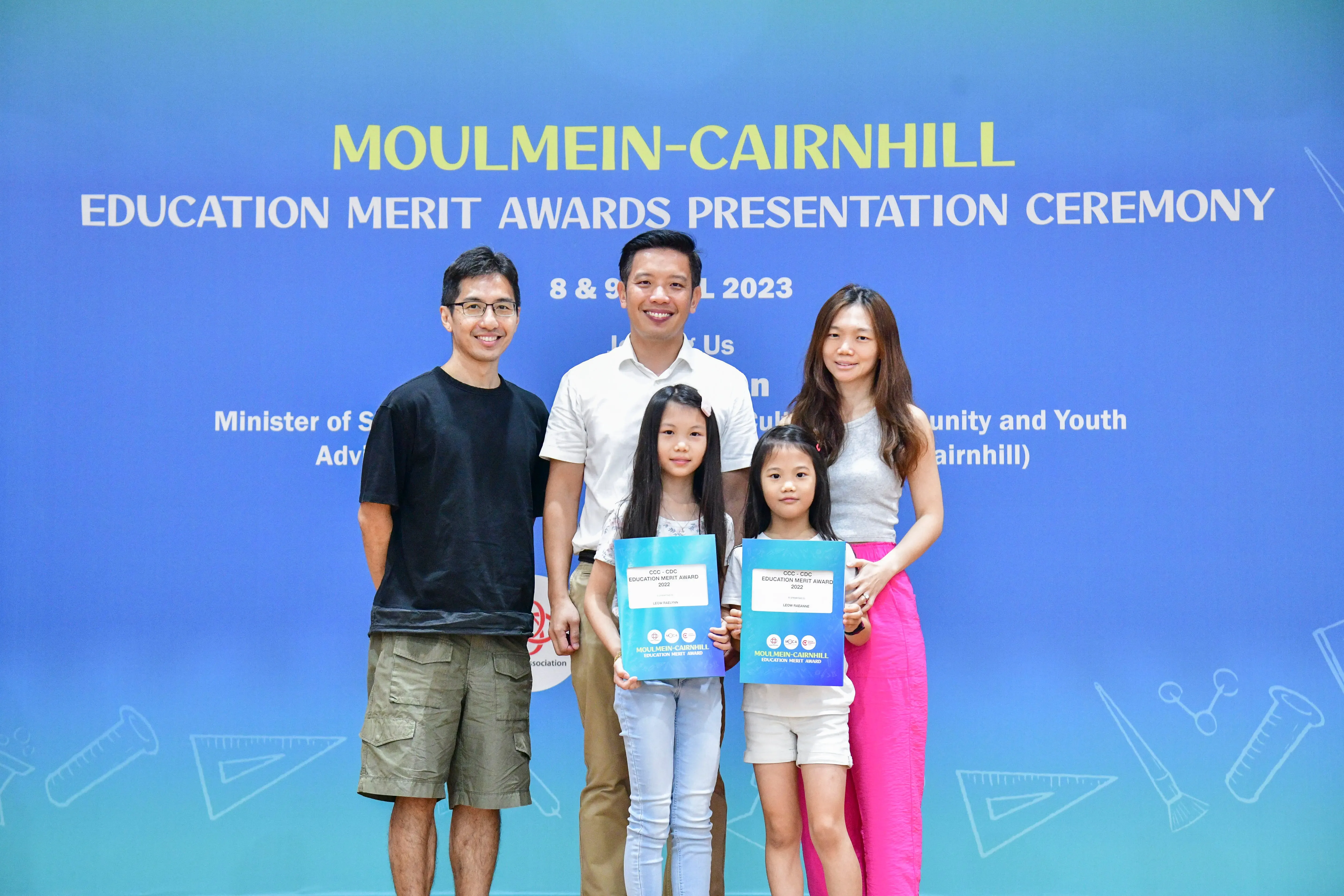 Two adults and two girls holding award certificates pose before a Moulmein-Cairnhill Education Merit Awards backdrop.