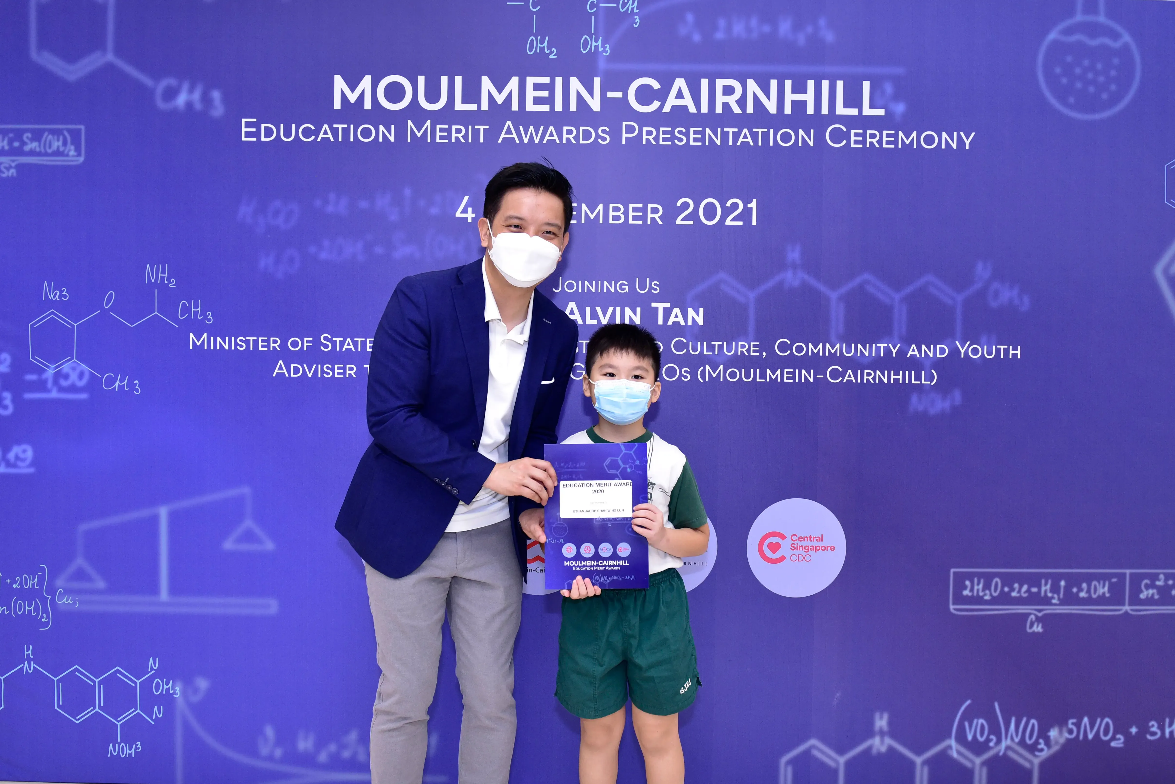 A masked man and young boy hold an award certificate at the Moulmein-Cairnhill Education Merit Awards 2021 ceremony.