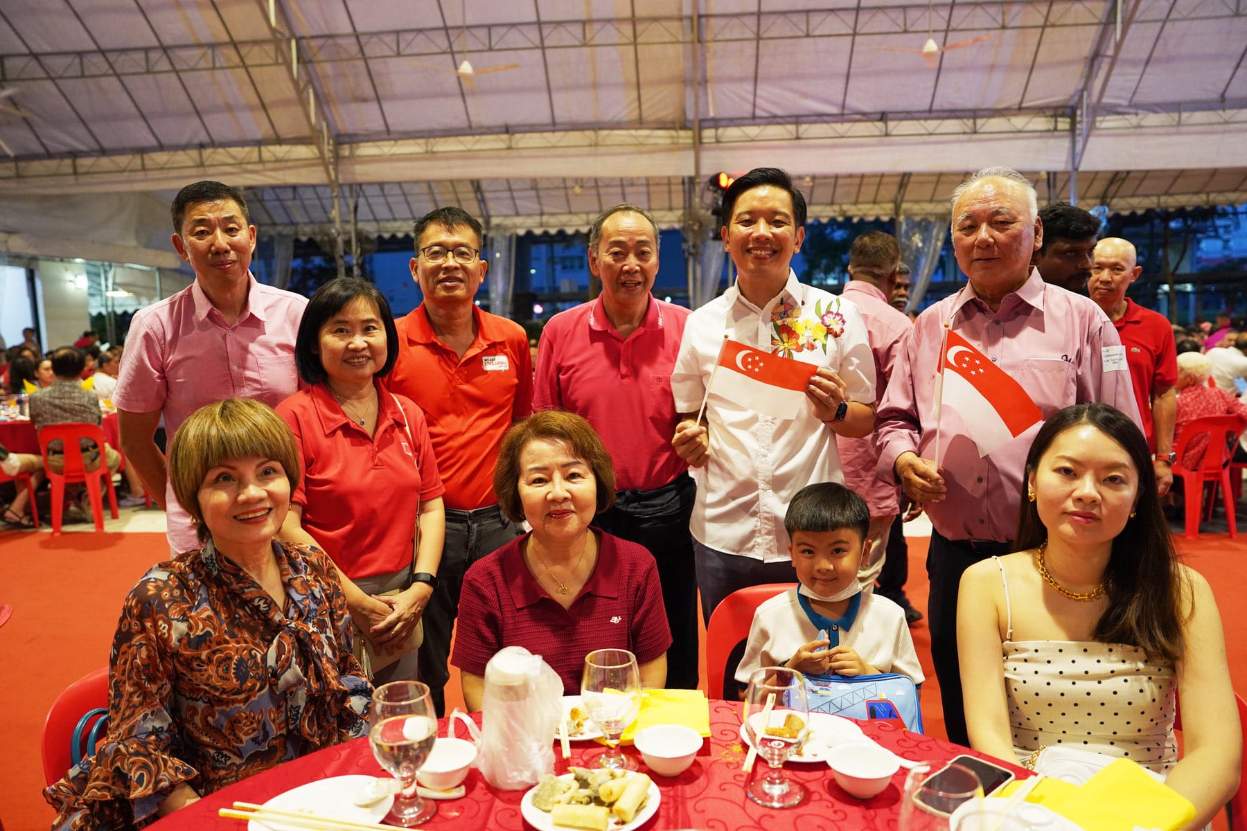 A group of people pose at a red-themed outdoor dinner event, with two holding small Singapore flags.