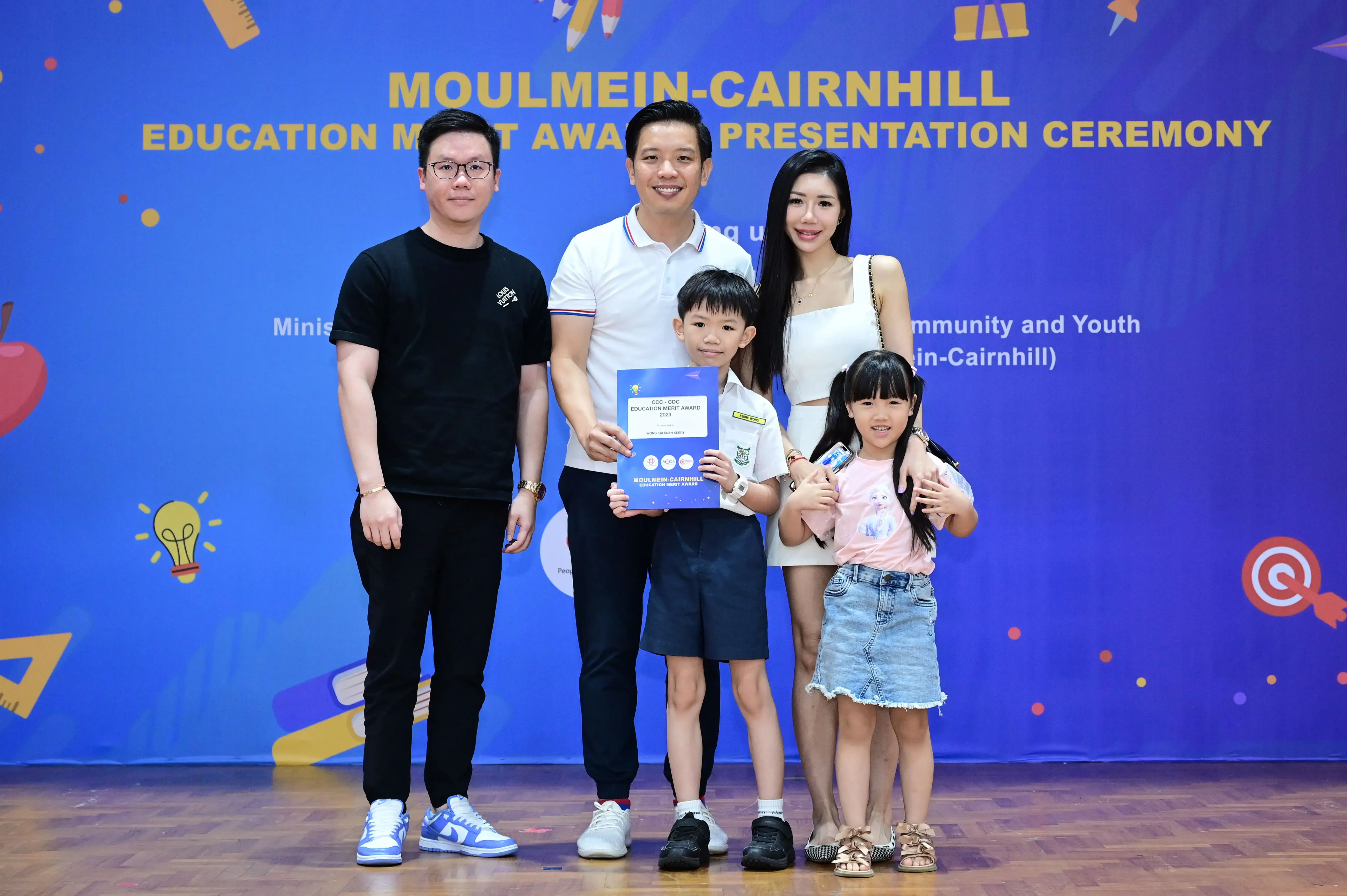 Five people pose before a Moulmein-Cairnhill Education Merit Award Presentation Ceremony banner, a boy holding a certificate.
