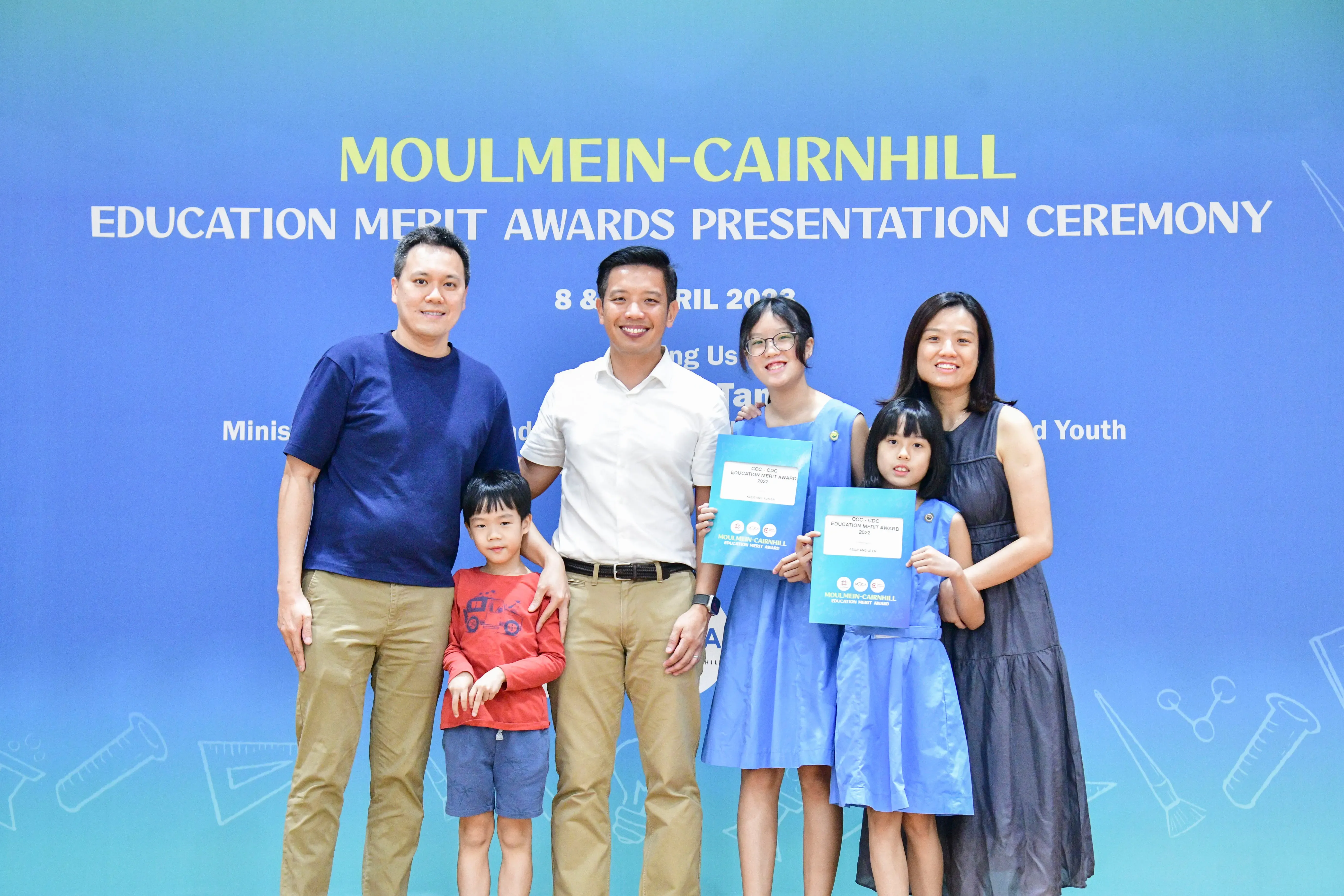 Six people pose in front of a blue Moulmein-Cairnhill Education Merit Awards Presentation Ceremony banner, two girls holding award certificates.