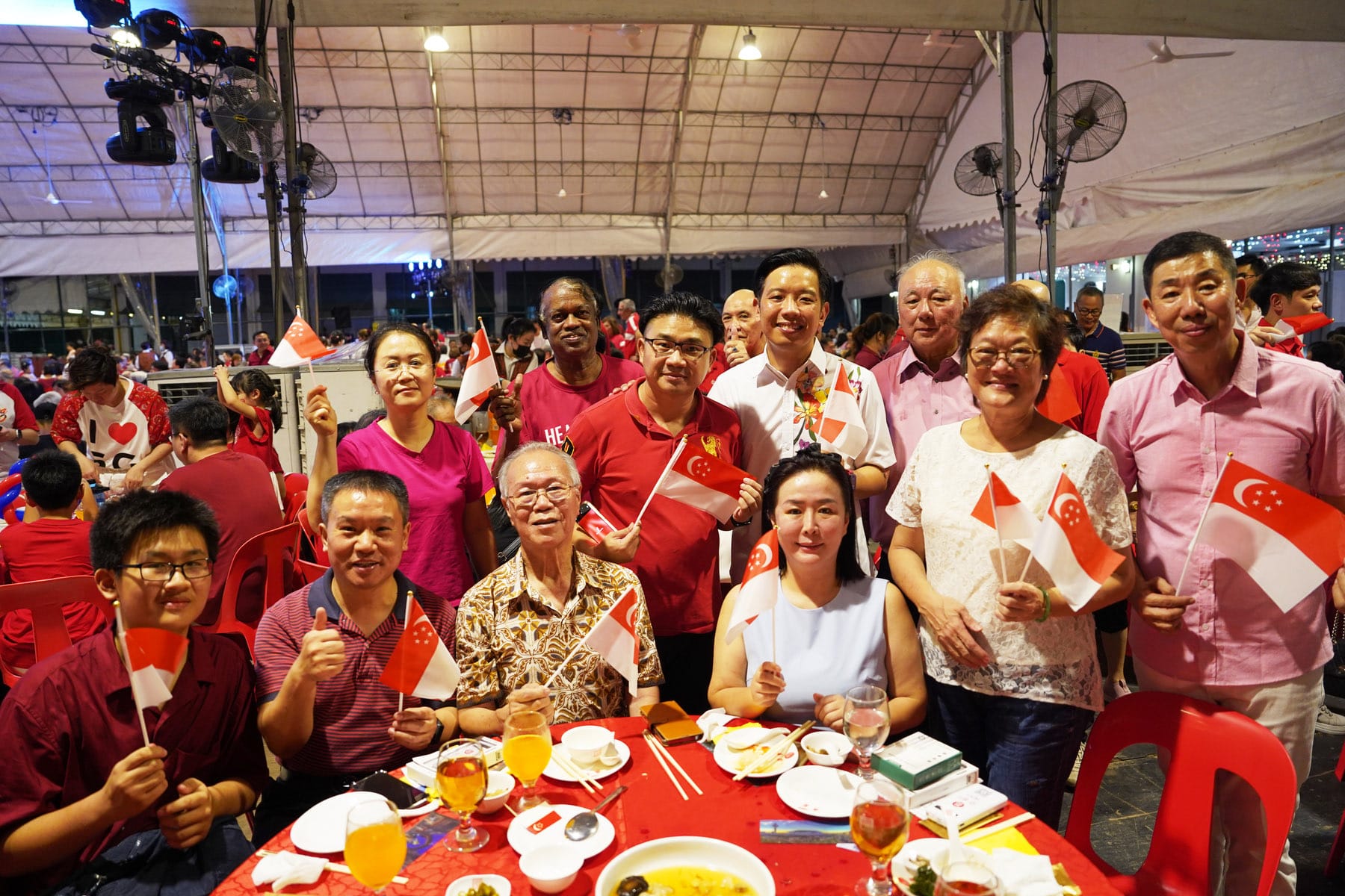A diverse group of smiling people waving small Singapore flags at a festive outdoor dinner under a tent.