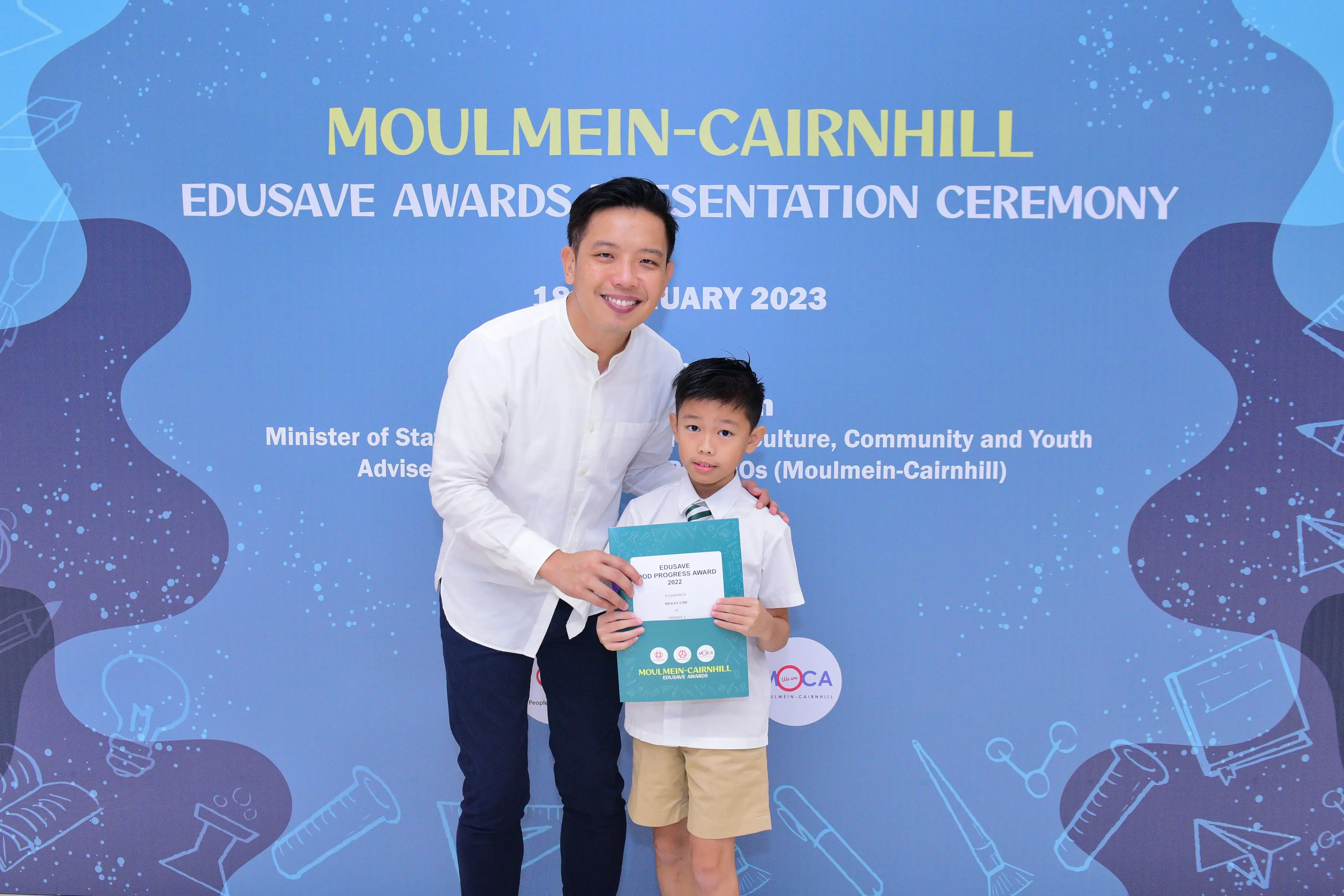 A man and a young boy pose together holding an Edusave award certificate at the Moulmein-Cairnhill Awards Ceremony 2023.