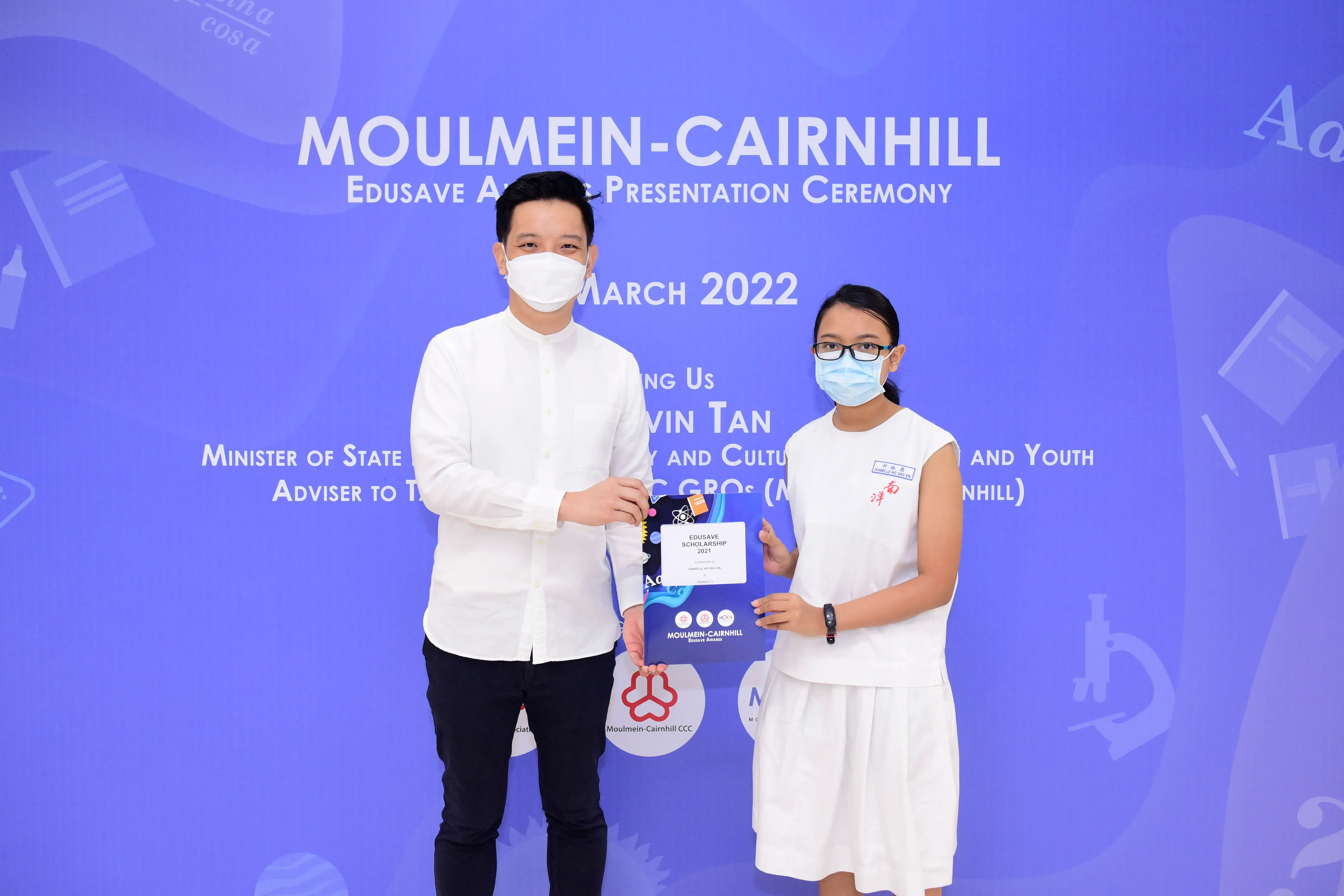 Two masked people hold an Edusave Scholarship certificate at the Moulmein-Cairnhill Award Ceremony, March 2022.