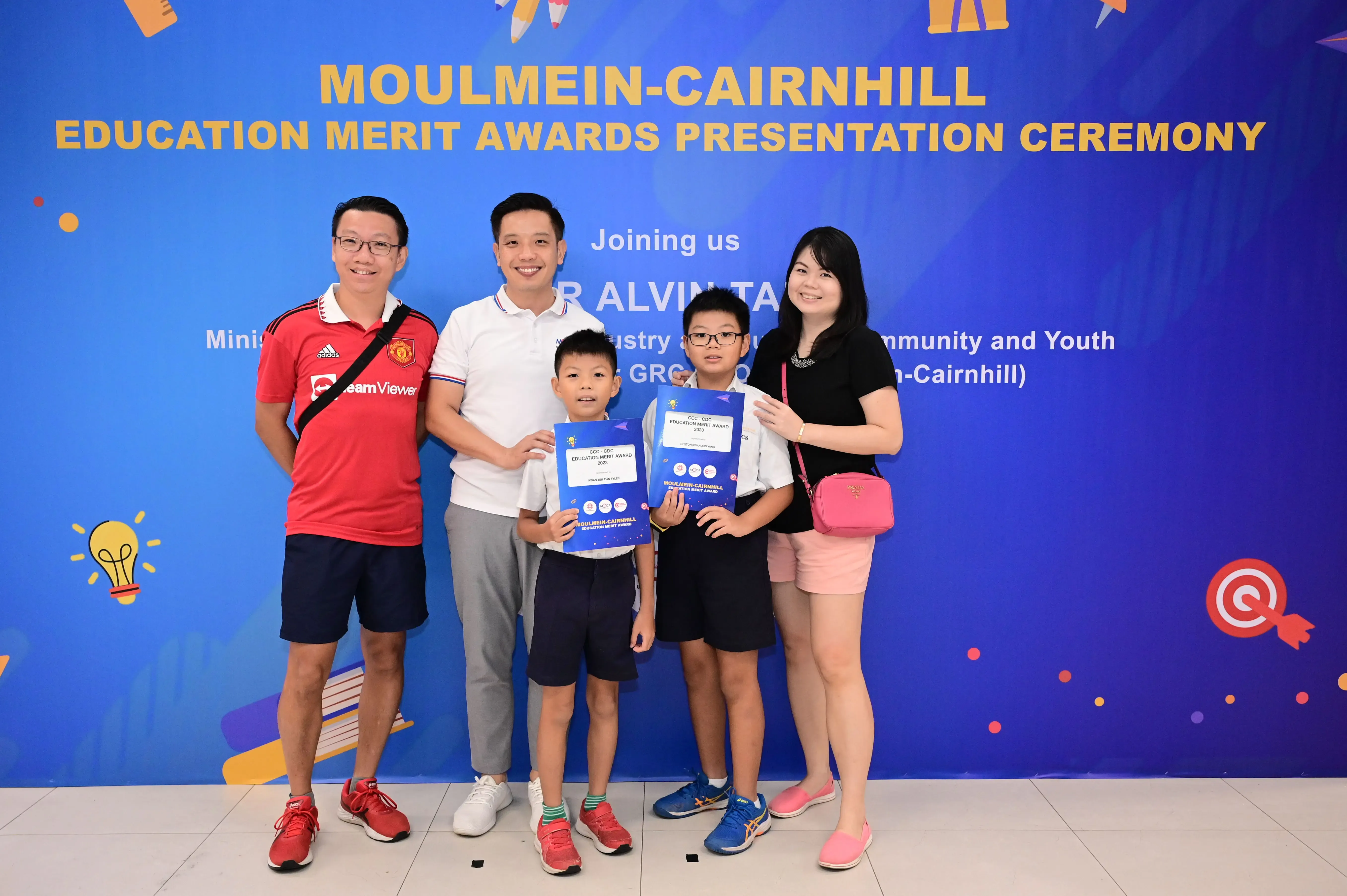 Five people pose before a Moulmein-Cairnhill Education Merit Awards banner, two boys holding blue award certificates.