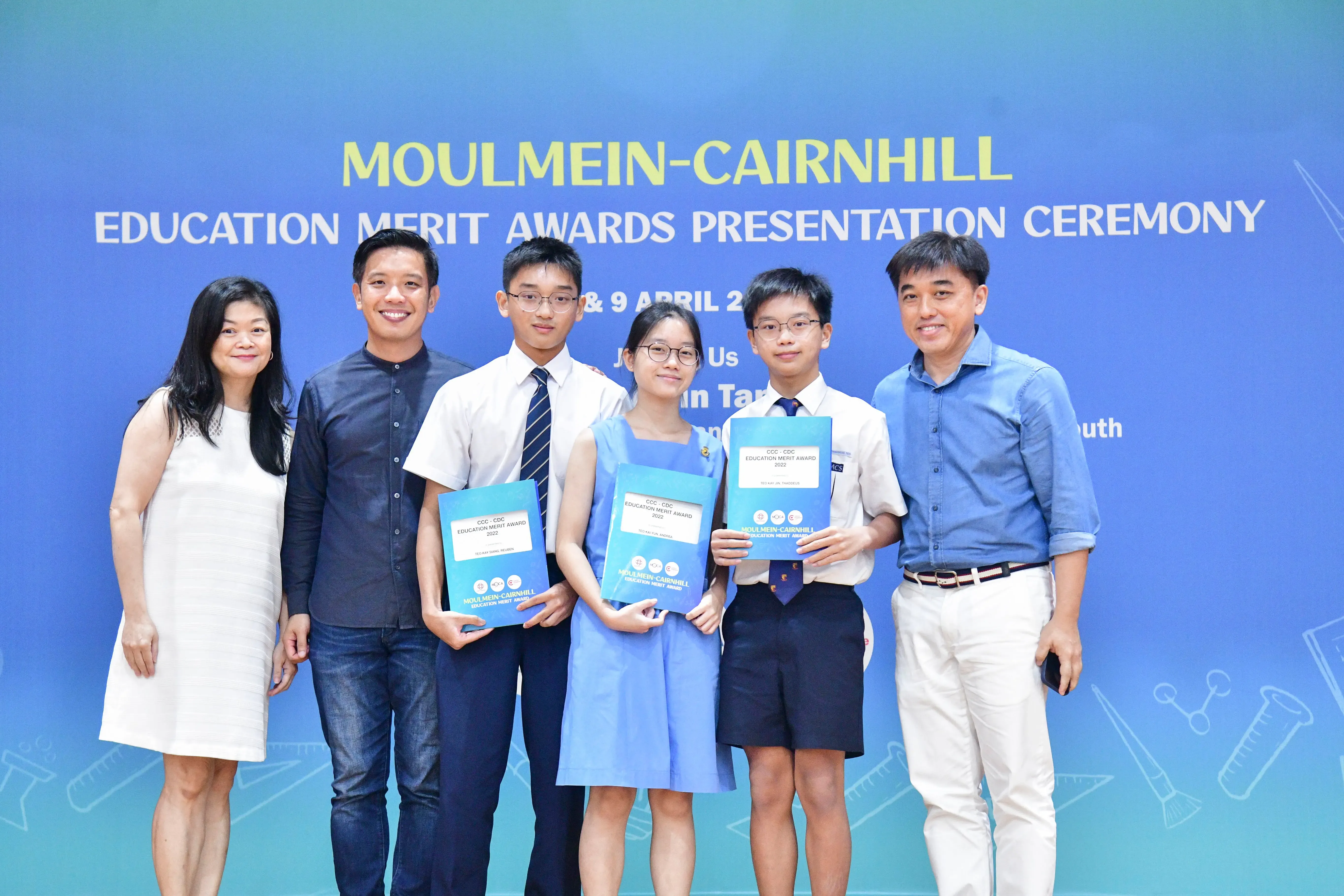 Six people pose in front of a blue Moulmein-Cairnhill Education Merit Awards Presentation Ceremony banner.