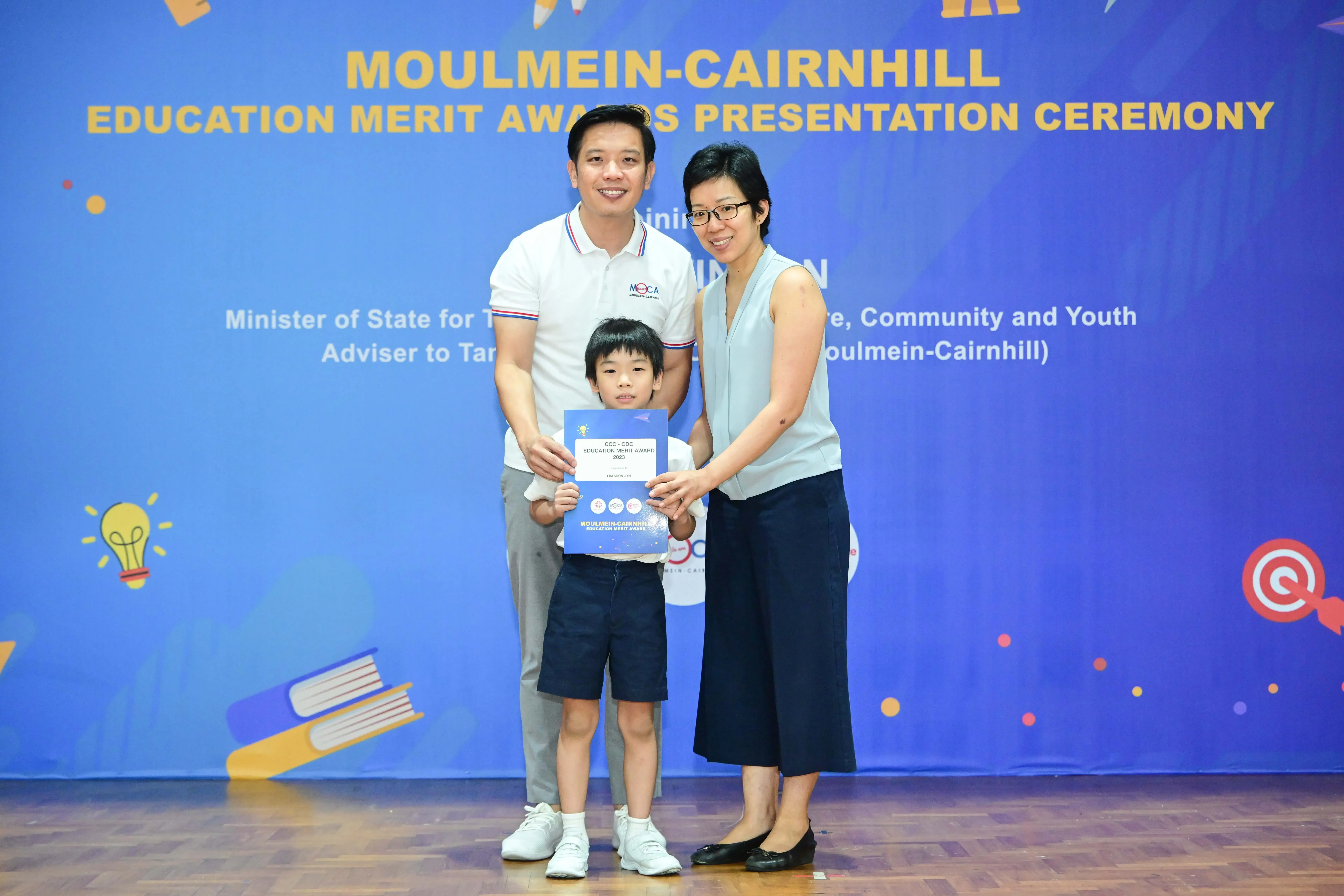 Two adults and a young boy hold an award certificate at the Moulmein-Cairnhill Education Merit Awards Presentation Ceremony.