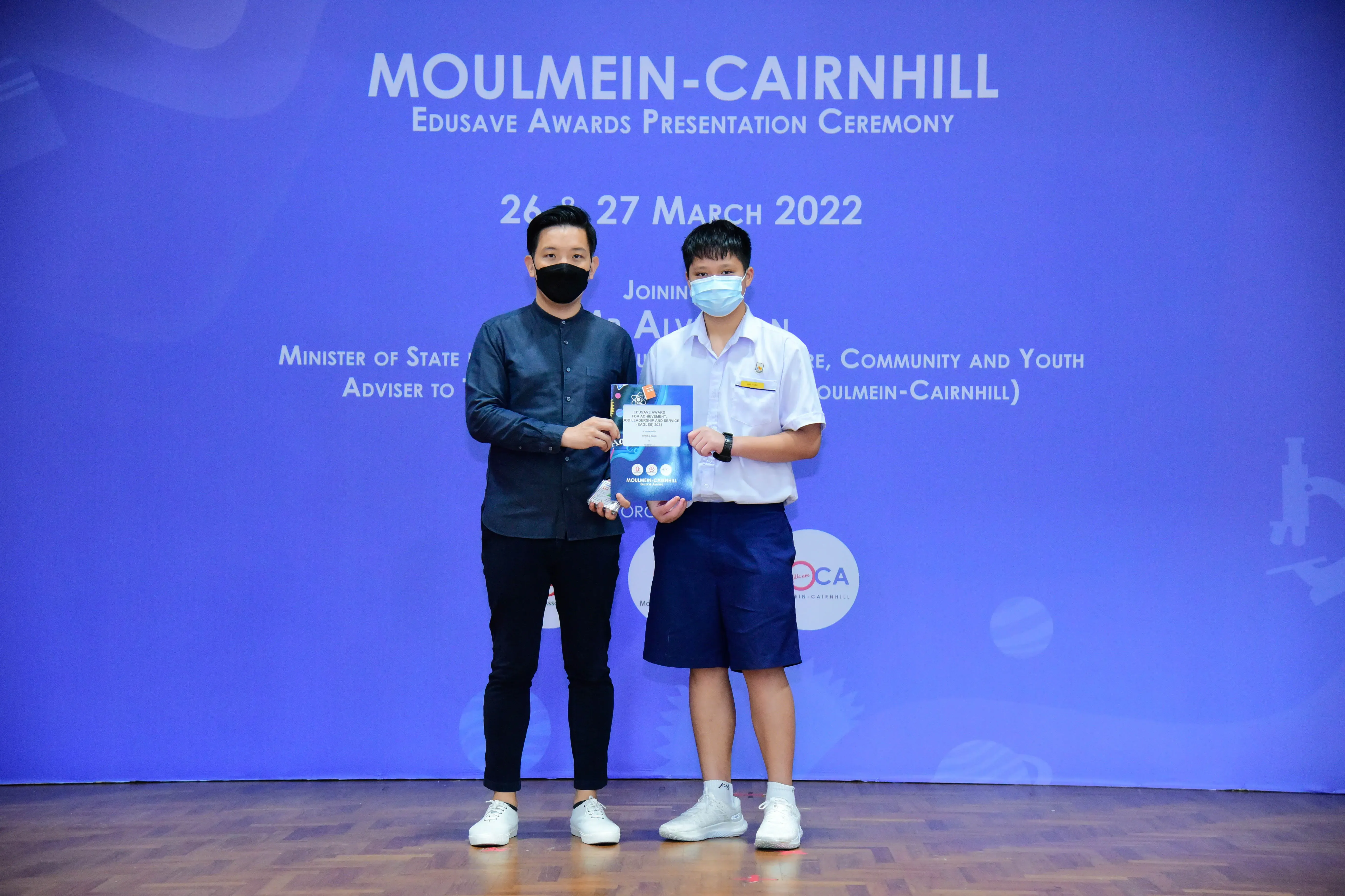 Two masked individuals hold an award booklet at the Moulmein-Cairnhill Edusave Awards Ceremony, March 2022.