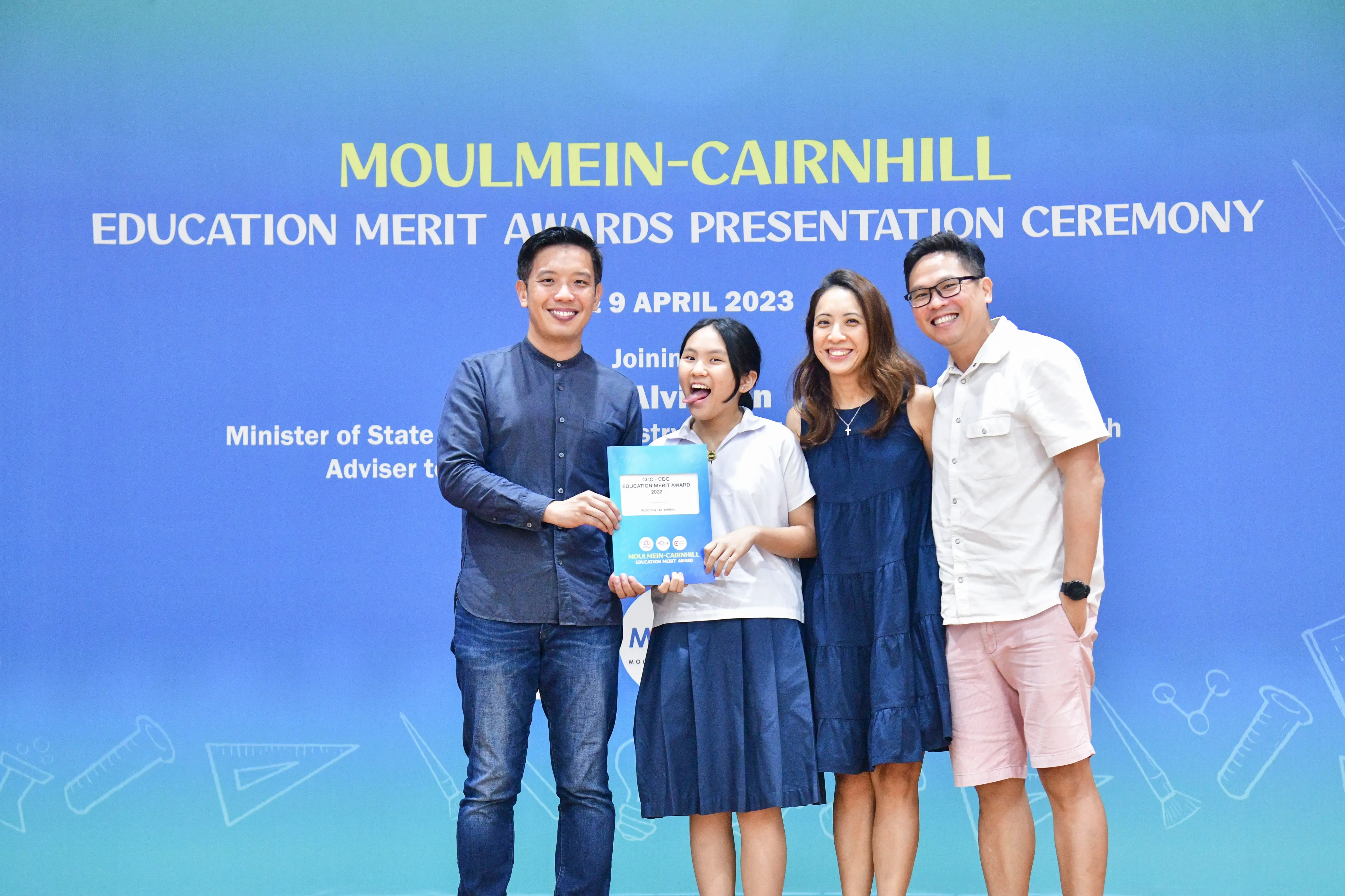 Four people smile before a Moulmein-Cairnhill Education Merit Awards banner dated 9 April 2023, holding a certificate.