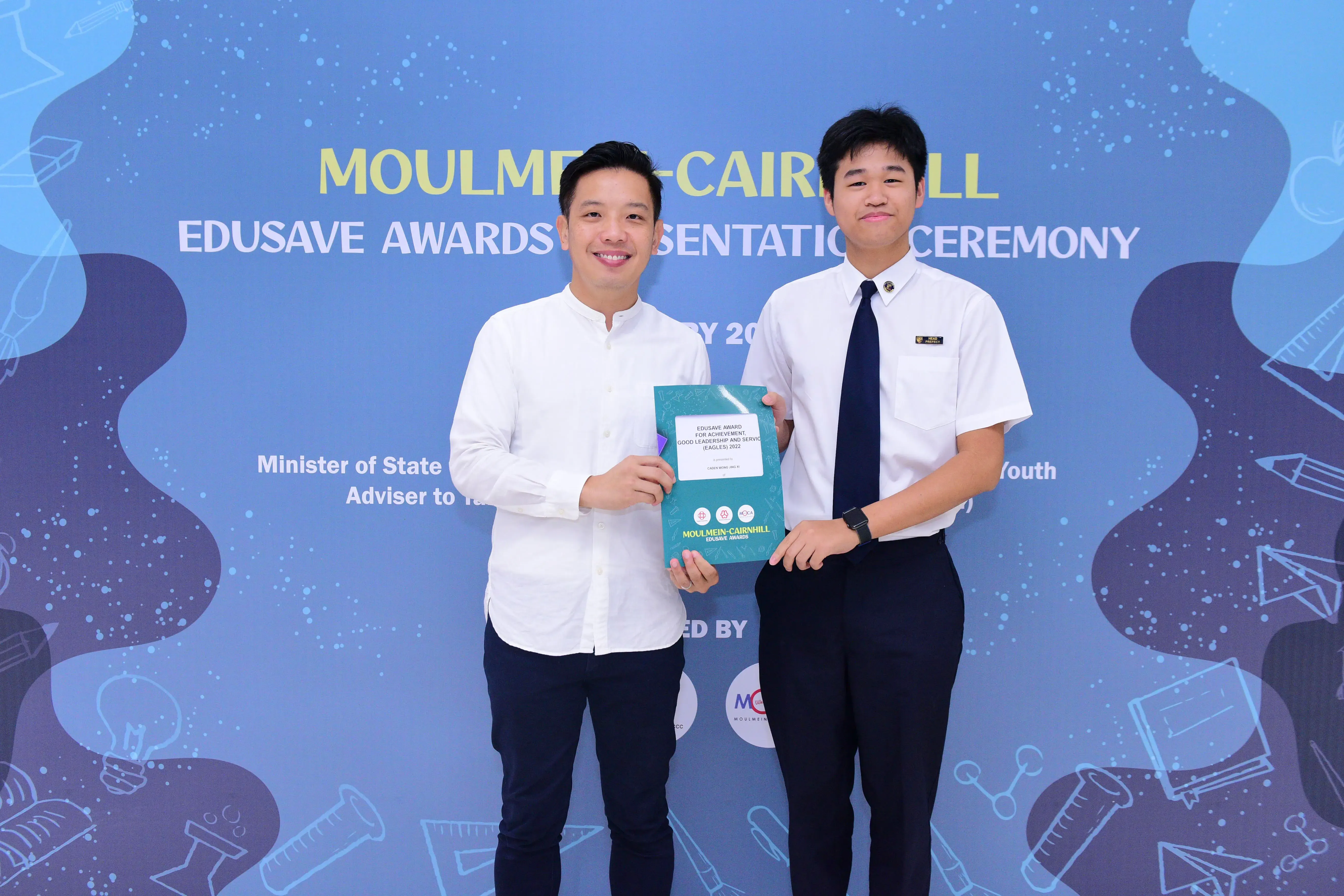 Two men smile holding a certificate at the Moulmein-Cairnhill Edusave Awards Presentation Ceremony backdrop.