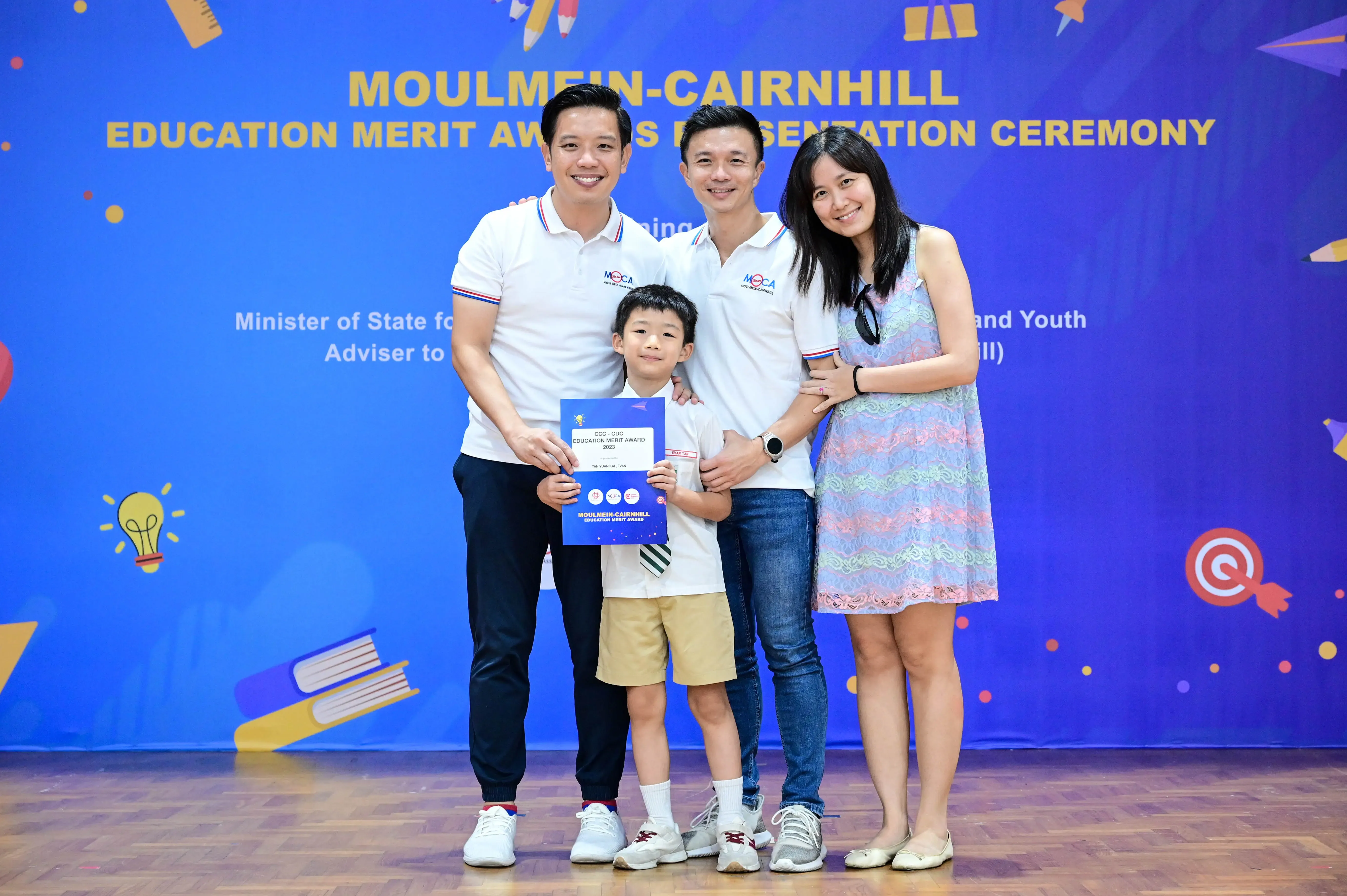 Three adults and a young boy pose smiling at the Moulmein-Cairnhill Education Merit Awards ceremony, boy holds a certificate.