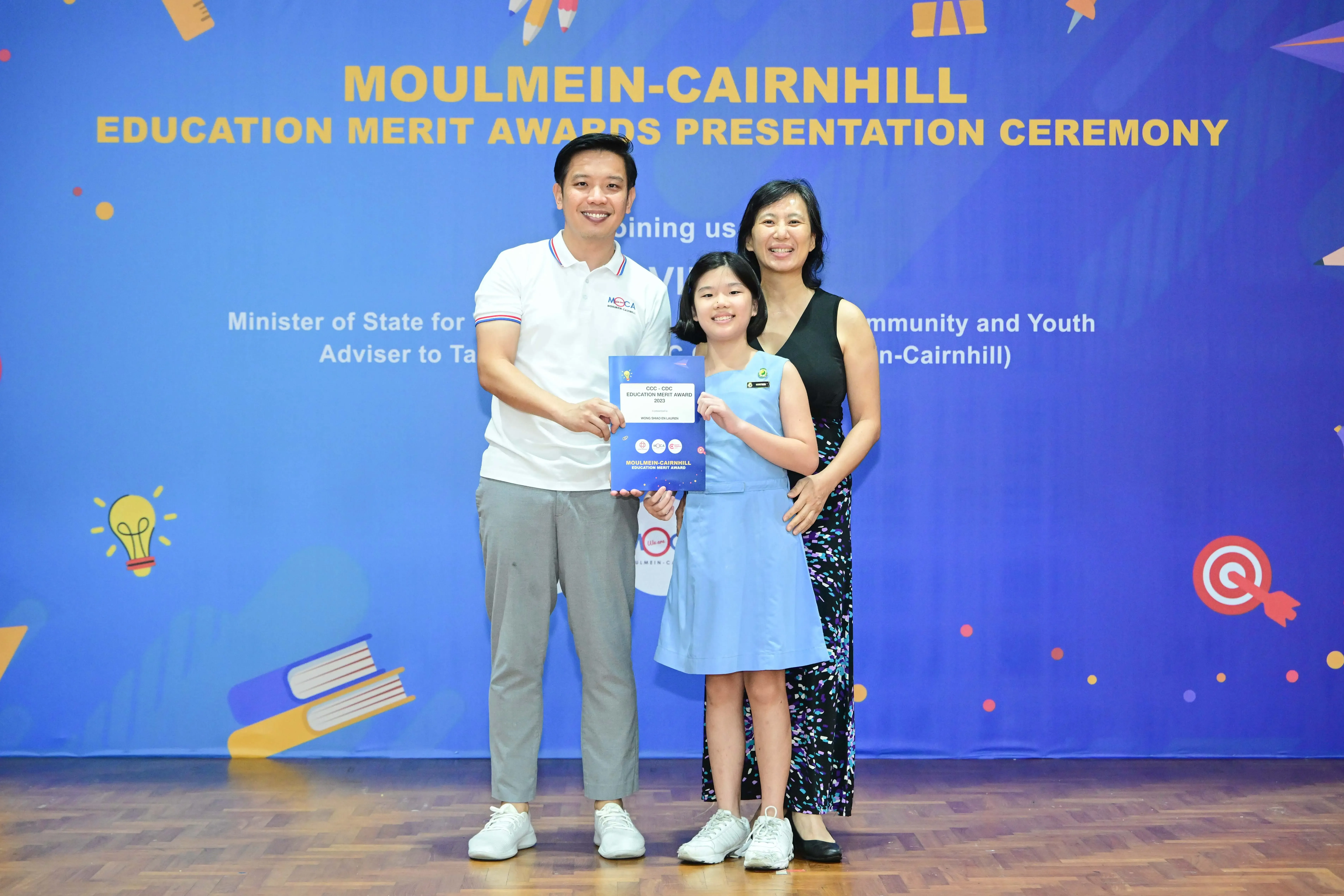 Two adults and a girl hold an award certificate at the Moulmein-Cairnhill Education Merit Awards Presentation Ceremony.