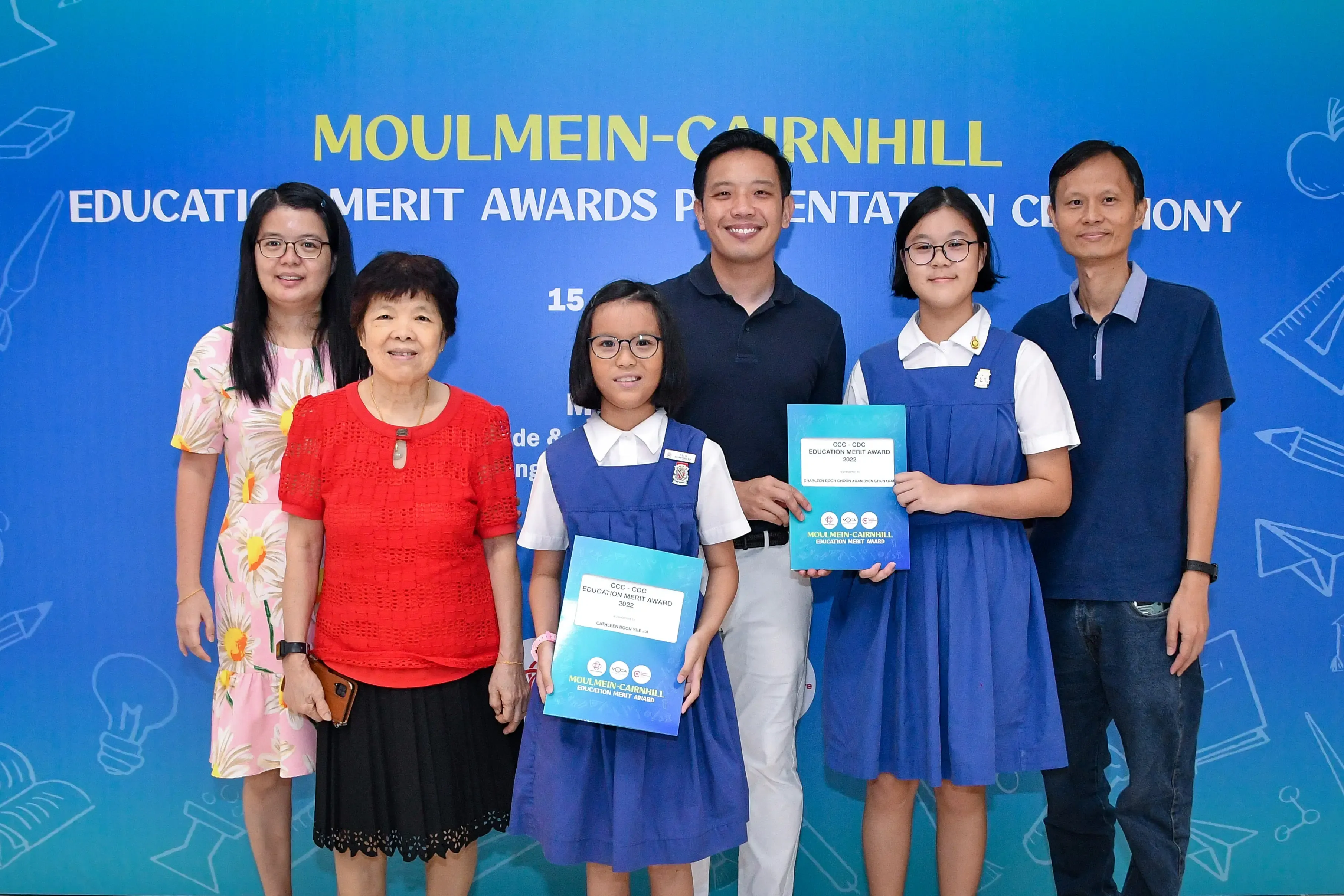 Six people pose in front of a Moulmein-Cairnhill Education Merit Awards banner, two girls holding award certificates.
