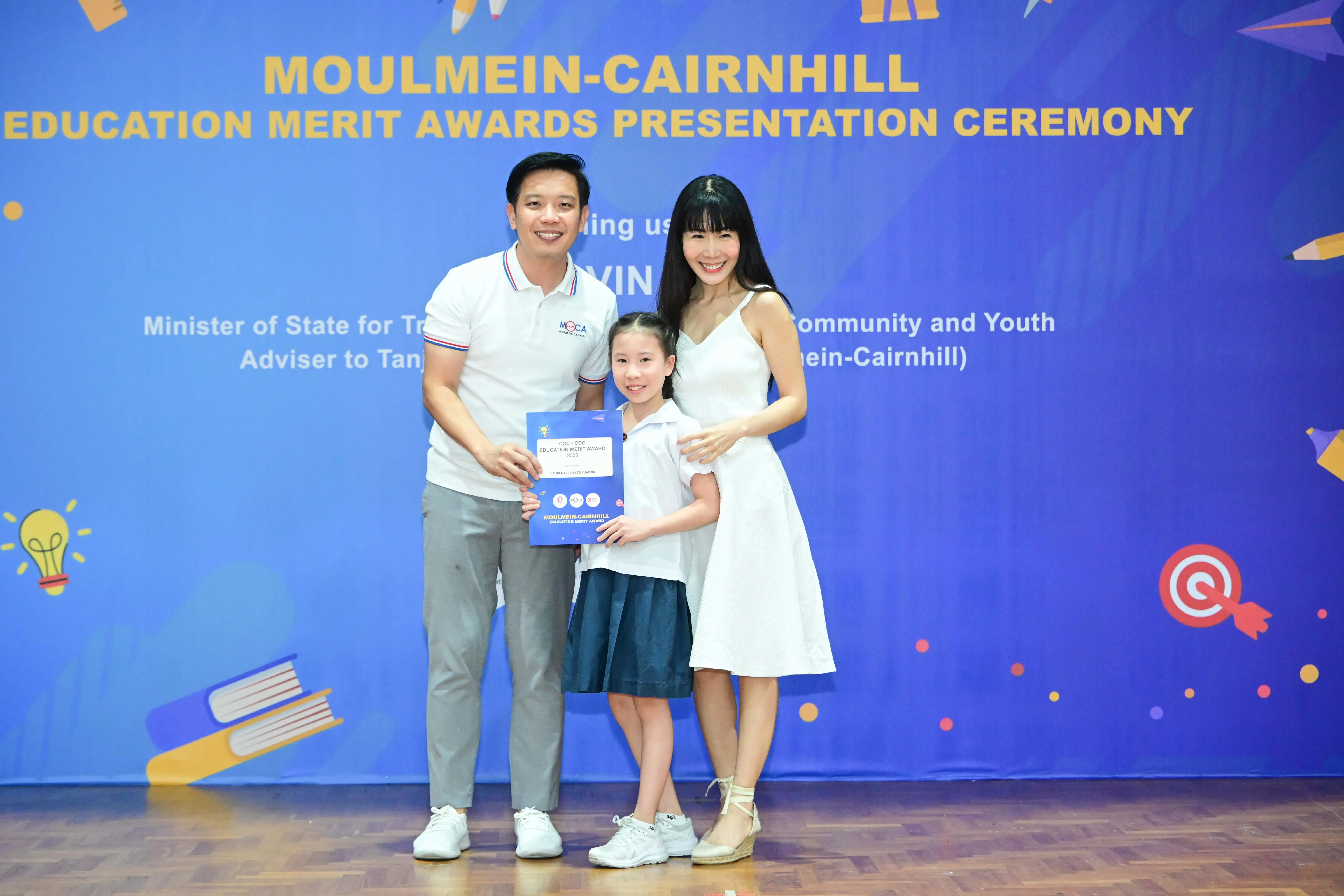 Two adults and a young girl holding an award certificate at the Moulmein-Cairnhill Education Merit Awards ceremony.