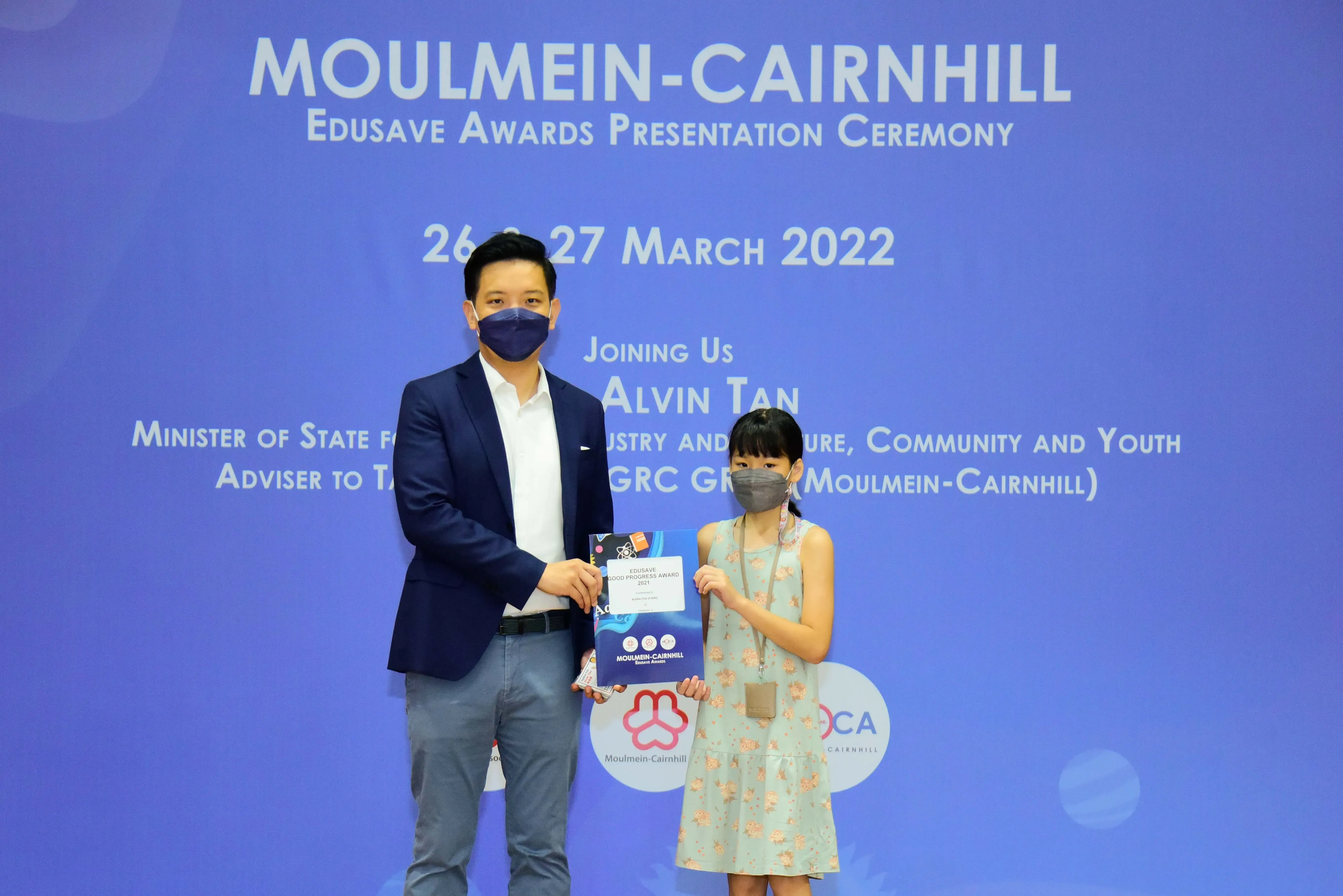 A masked man in a suit presents an Edusave award to a young masked girl at a March 2022 ceremony.