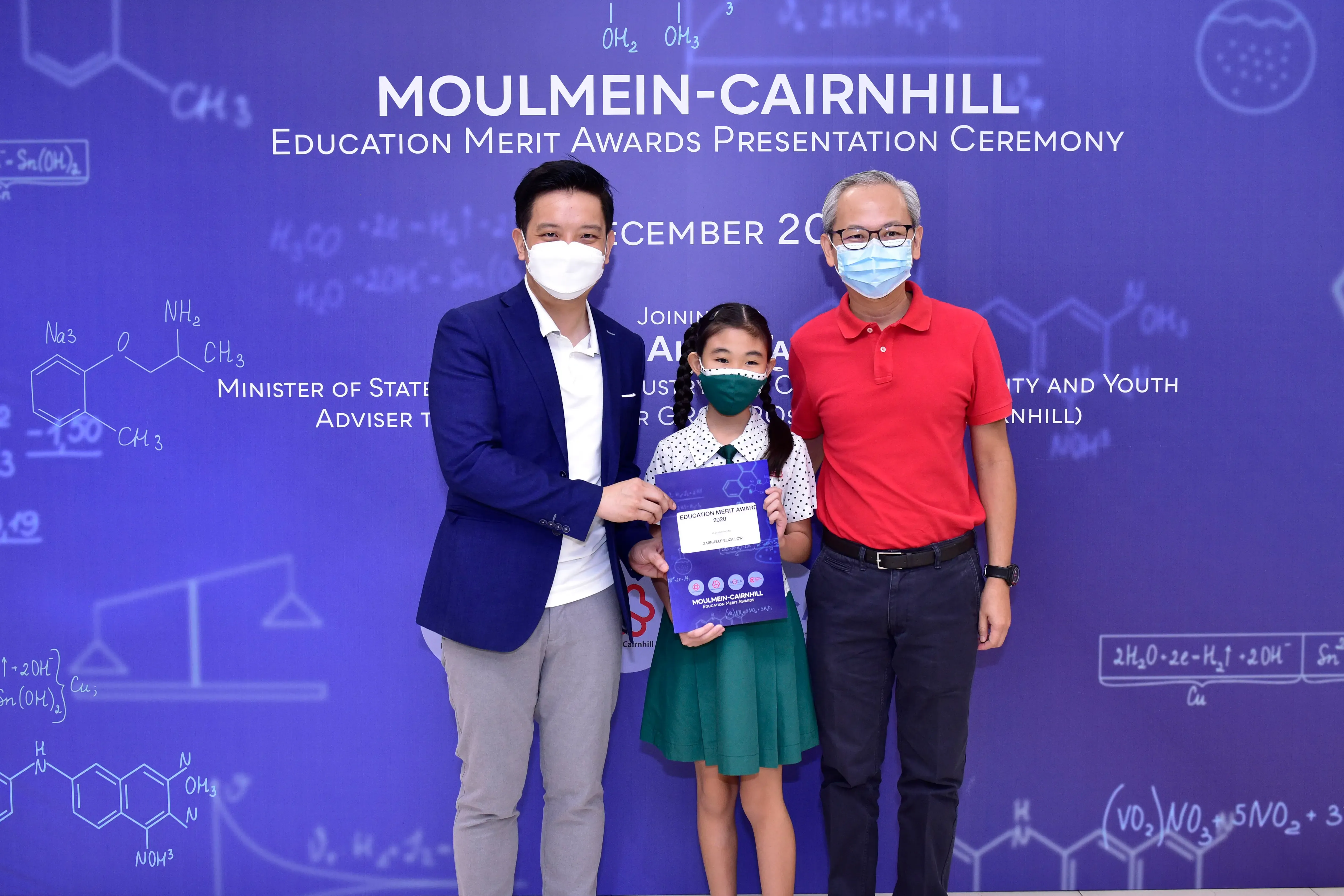 Two masked adults flank a young girl holding an award certificate at the Moulmein-Cairnhill Education Merit Awards ceremony.