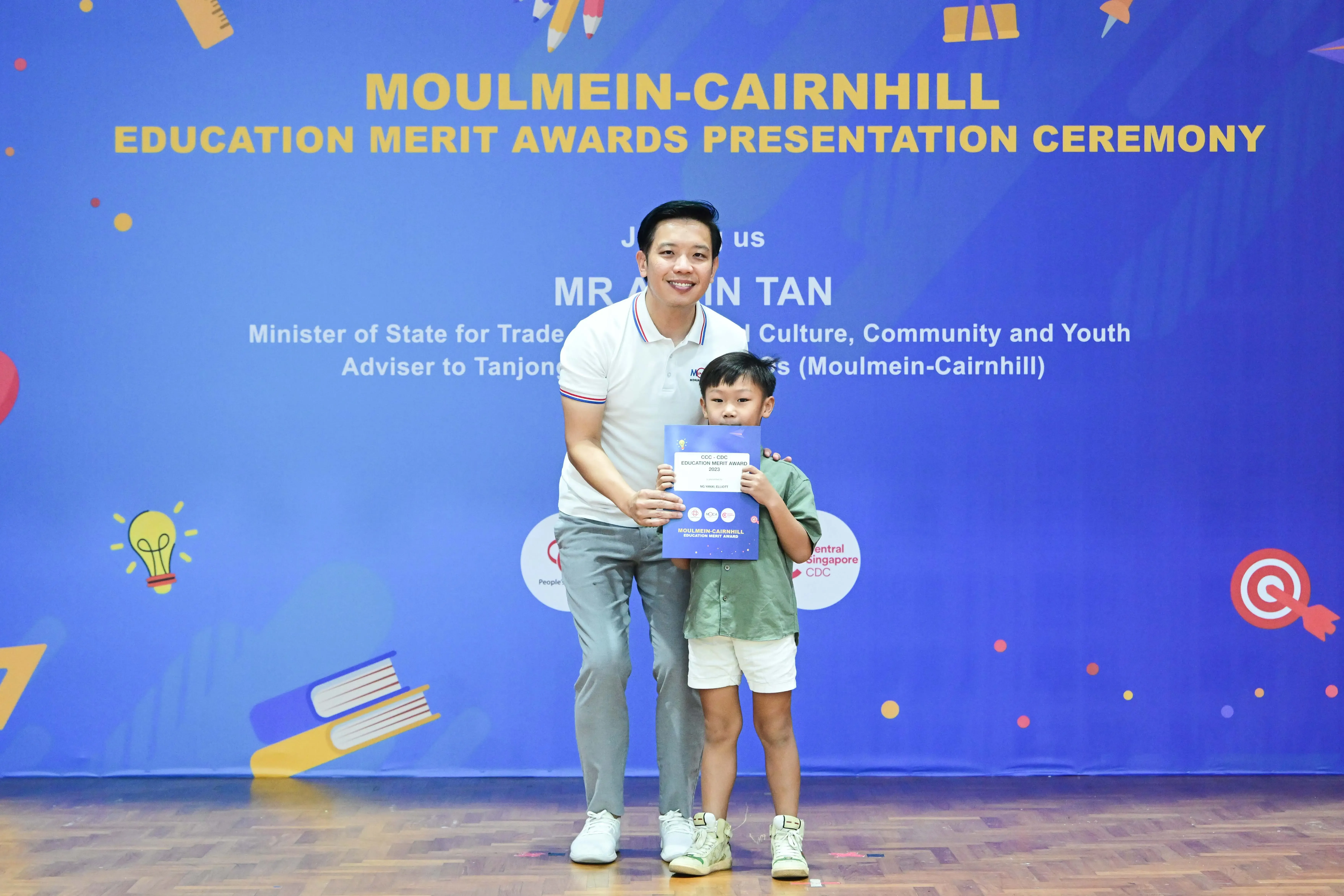 A man and a young boy hold a certificate in front of a Moulmein-Cairnhill Education Merit Awards banner.