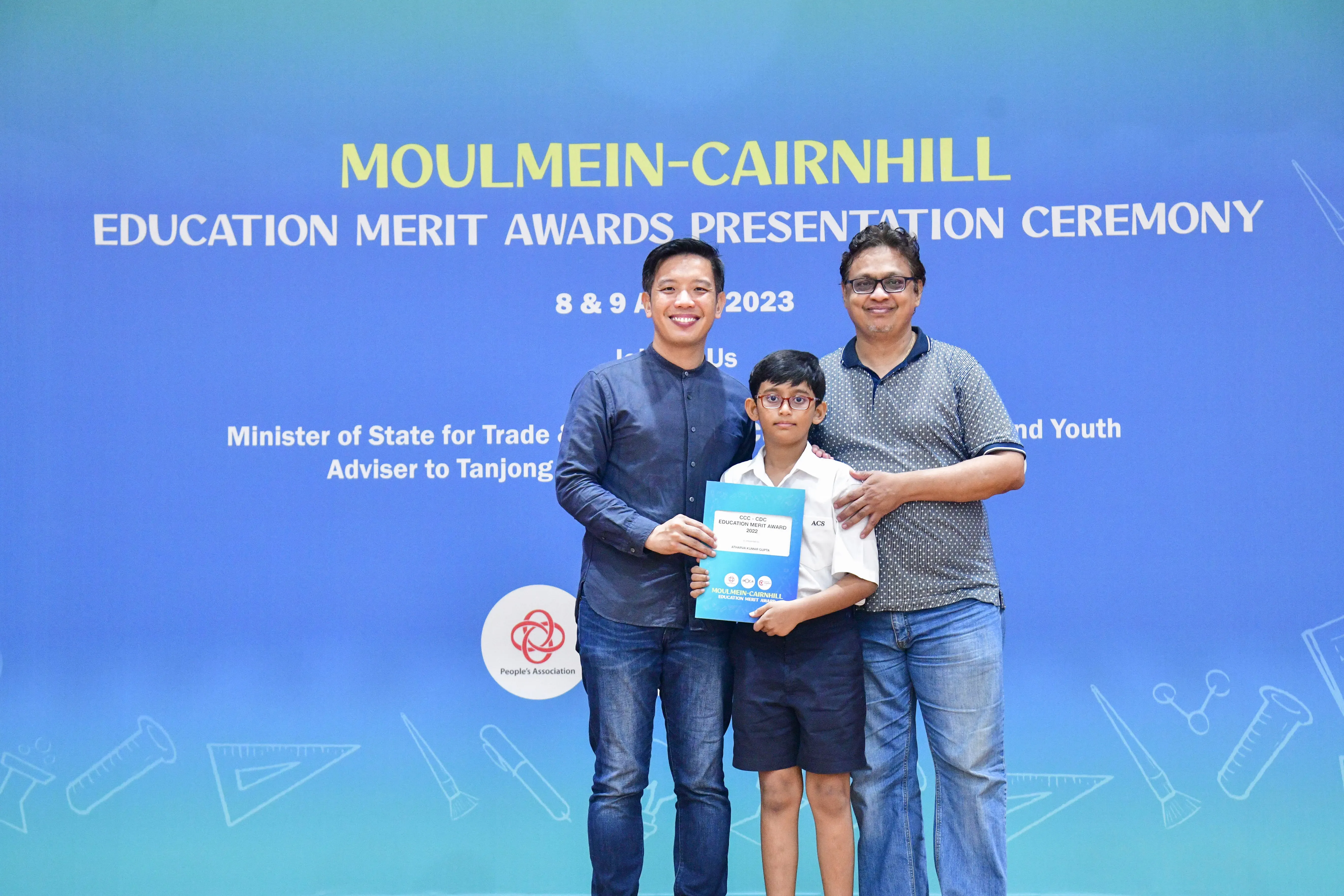 Two men and a boy holding an award certificate pose before a Moulmein-Cairnhill Education Merit Awards 2023 banner.