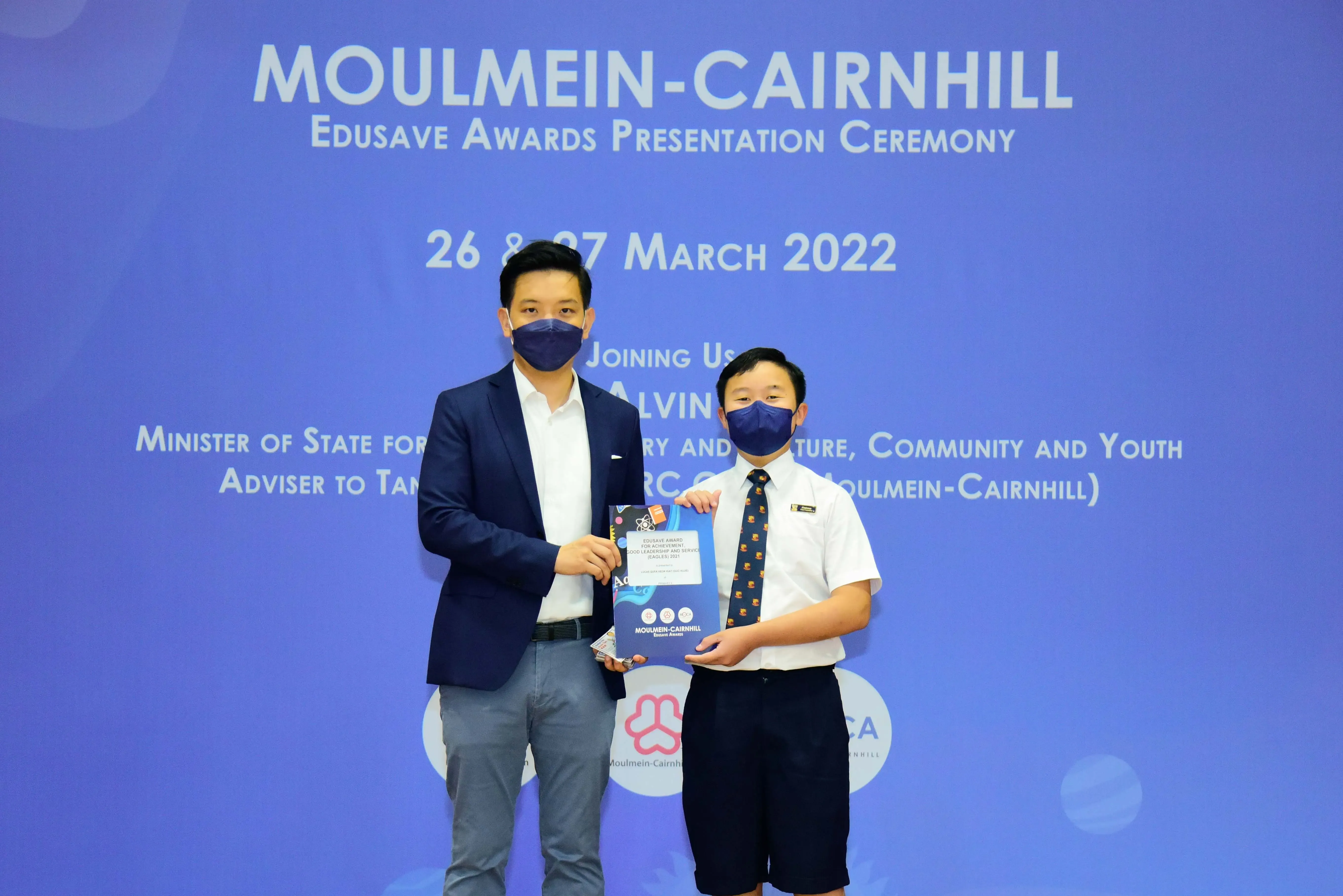 Two masked individuals hold an award at the Moulmein-Cairnhill Edusave Awards Presentation Ceremony, March 2022.
