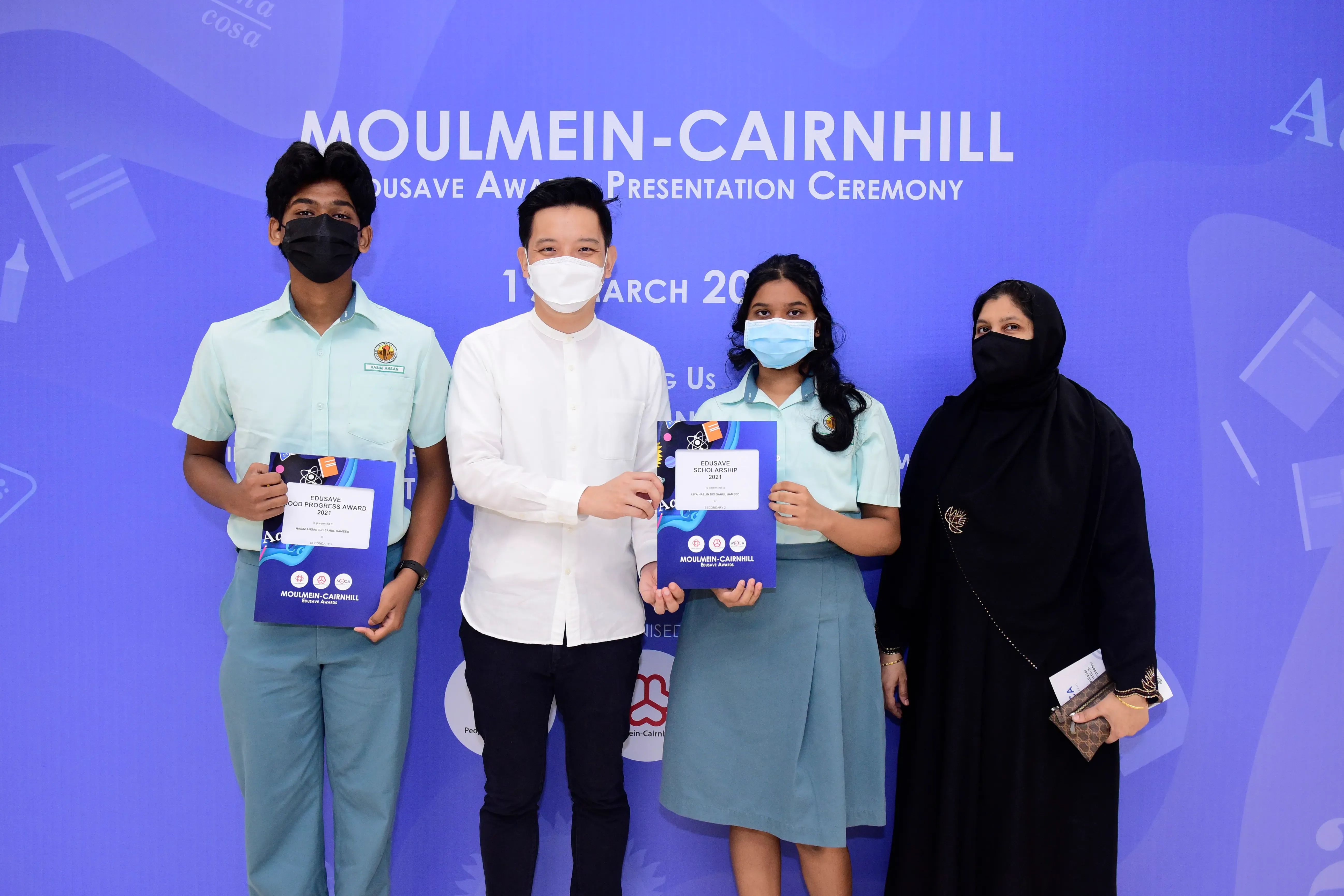 Four masked people pose at the Moulmein-Cairnhill Edusave Award Presentation Ceremony, two students holding award folders.
