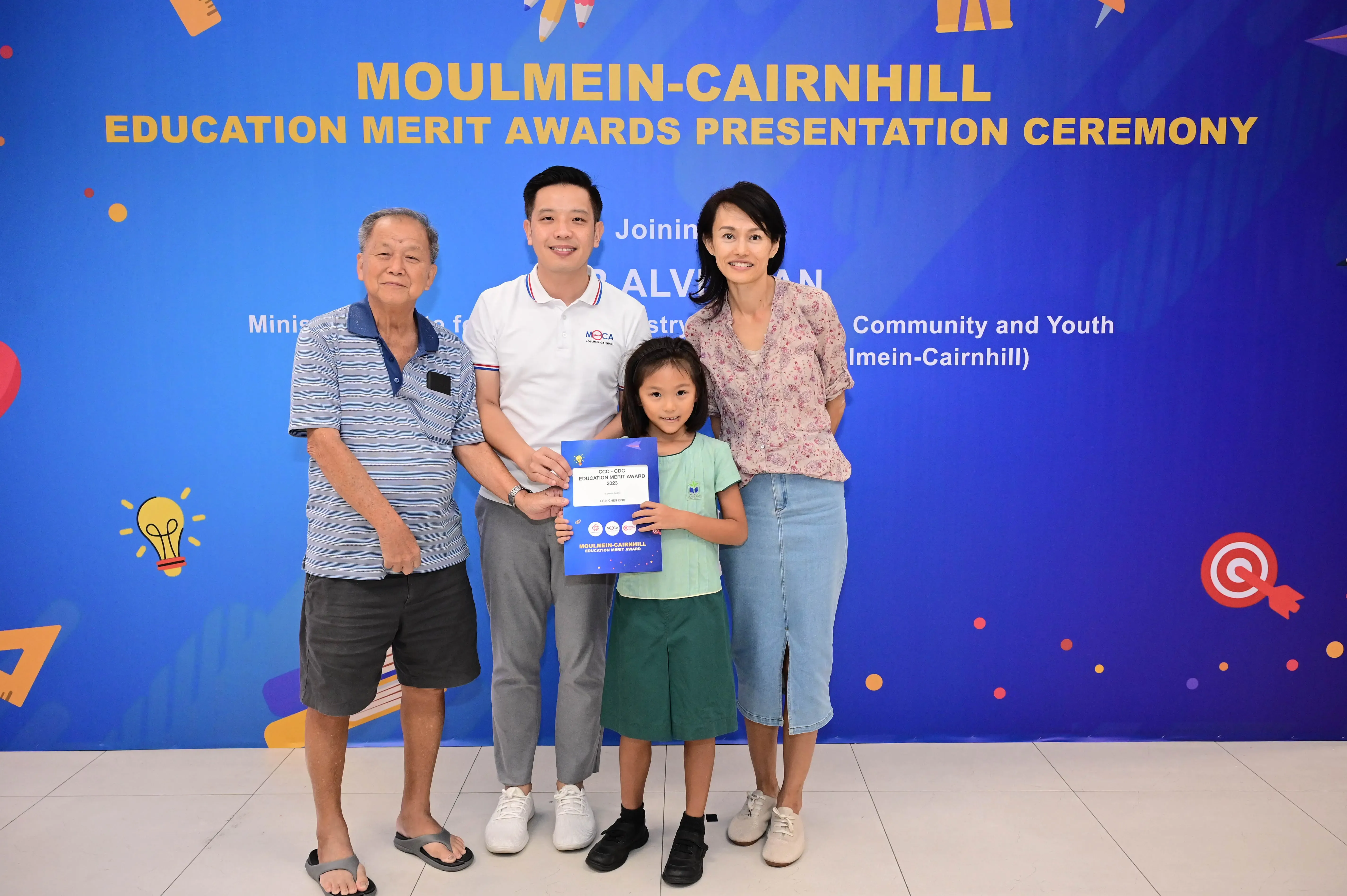 Four people pose before a Moulmein-Cairnhill Education Merit Awards banner, with a young girl holding a blue certificate.