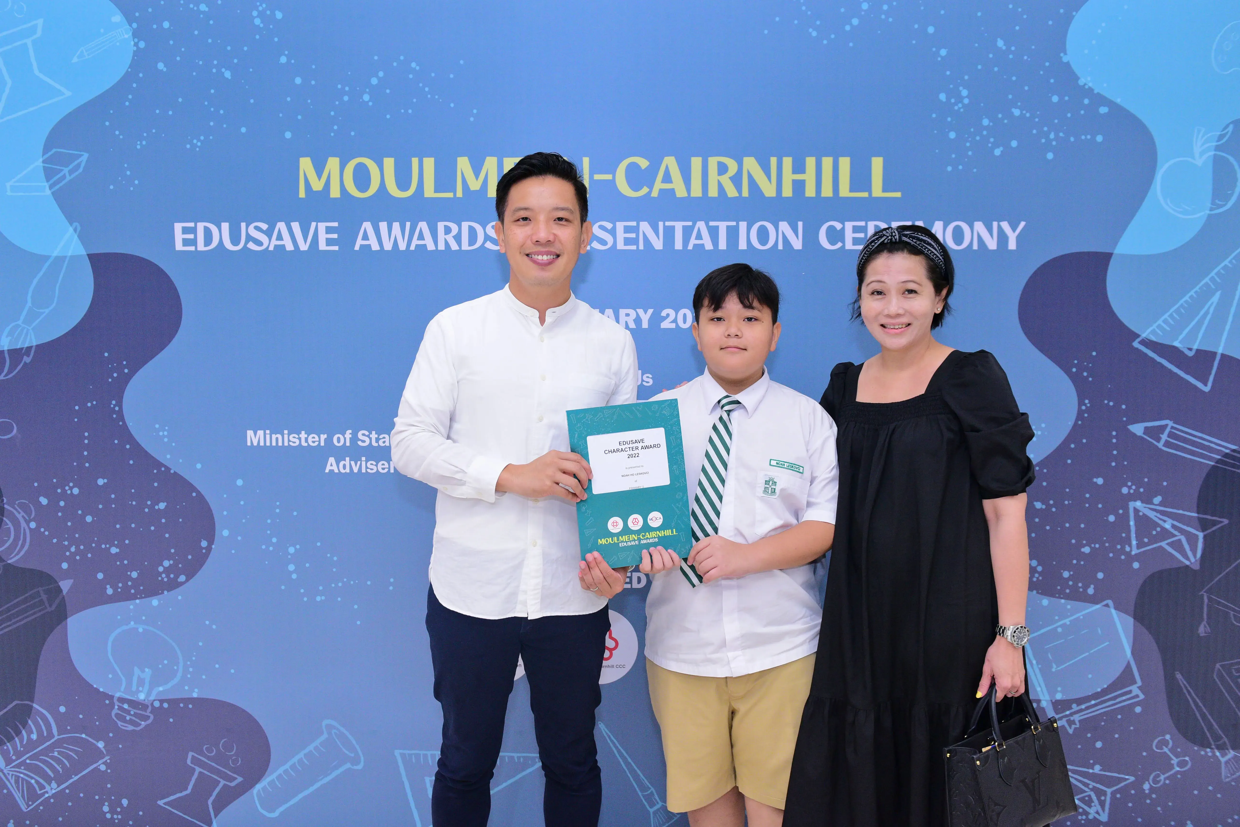 A boy in school uniform holds an Edusave award certificate flanked by two adults at Moulmein-Cairnhill ceremony.