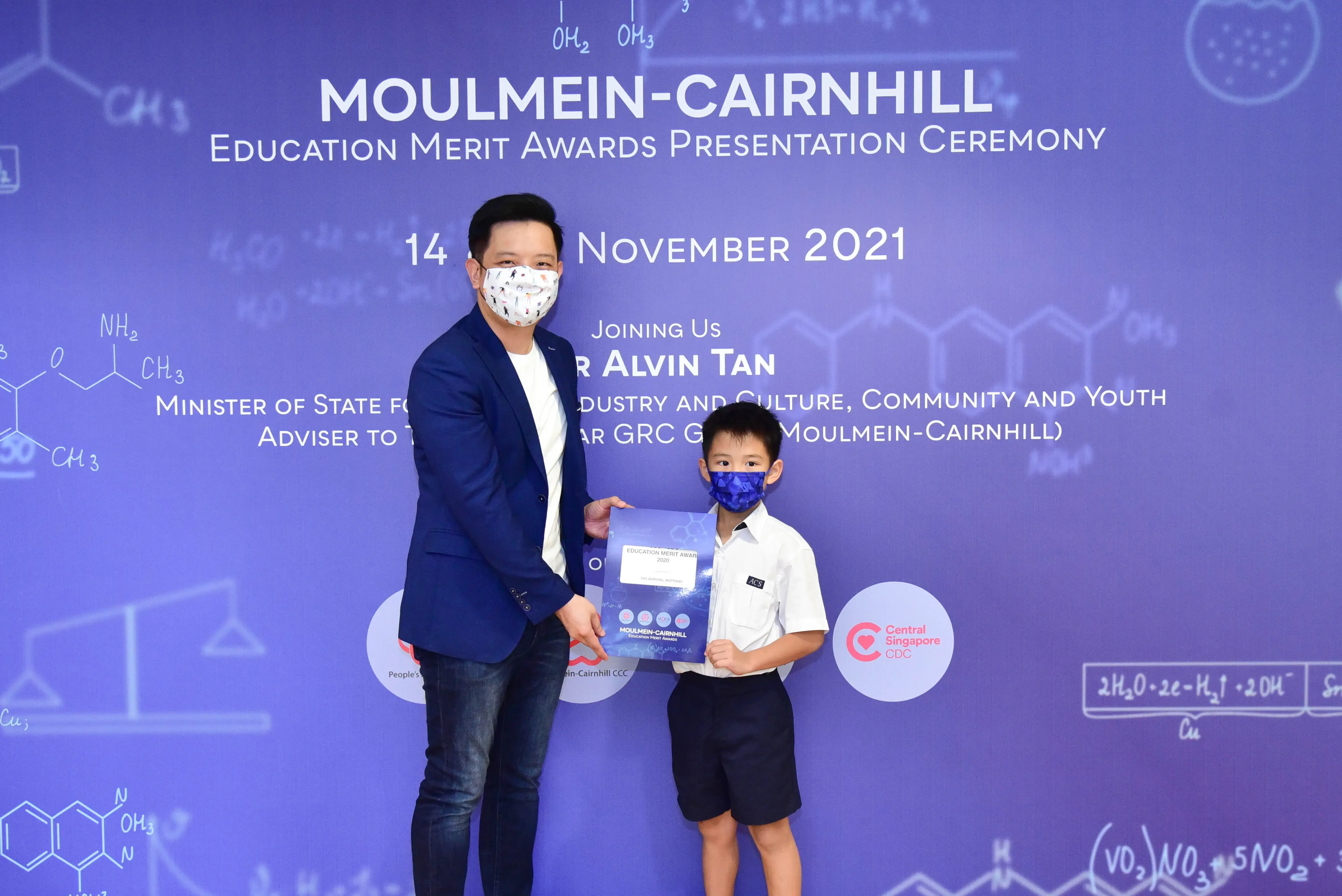 An adult and a young boy in school uniform hold a certificate at the Moulmein-Cairnhill Education Merit Awards 2021.