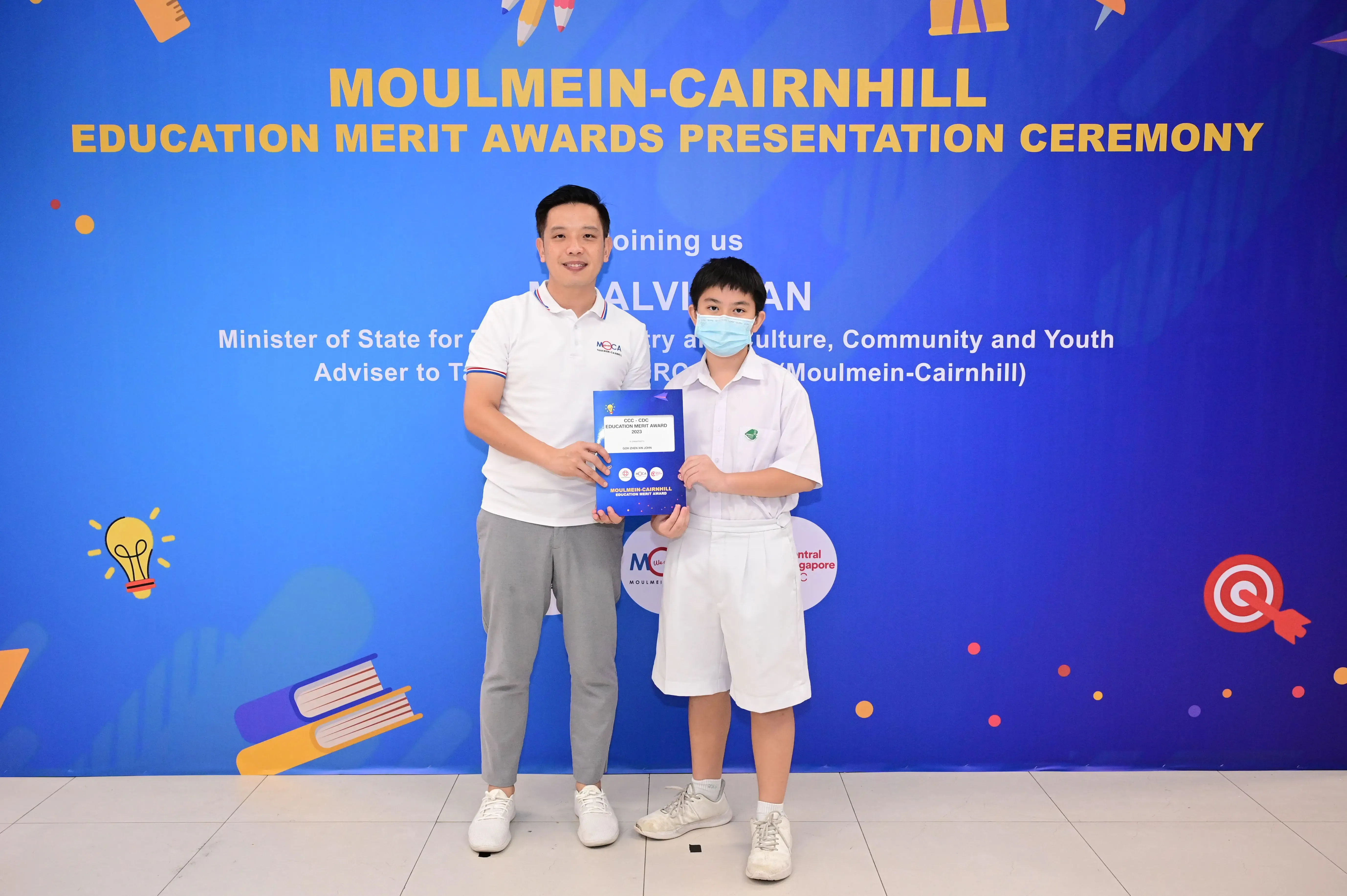 A man and a masked boy hold a blue award booklet at the Moulmein-Cairnhill Education Merit Awards ceremony.