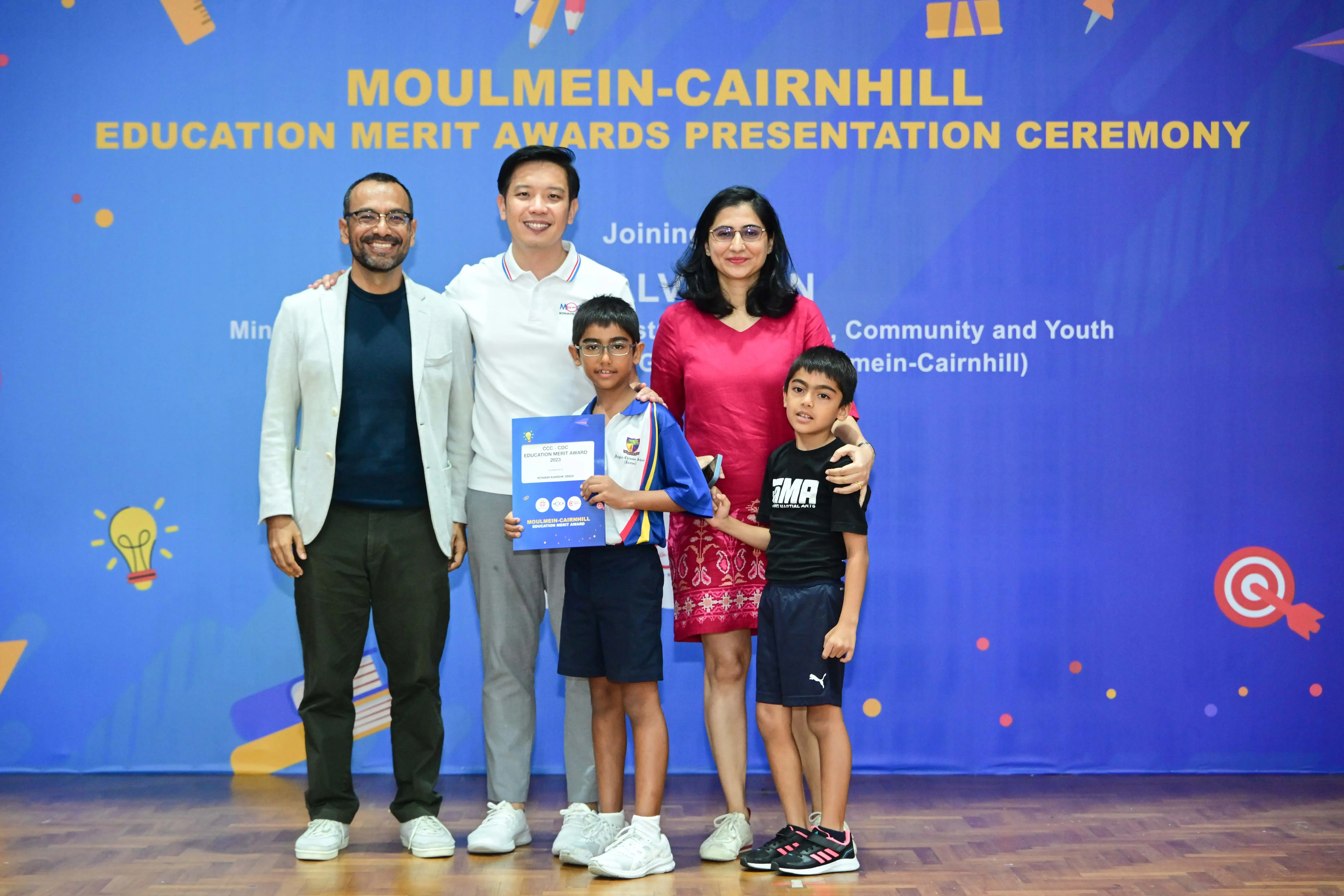 Five people pose at the Moulmein-Cairnhill Education Merit Awards Presentation Ceremony, with a boy holding a certificate.