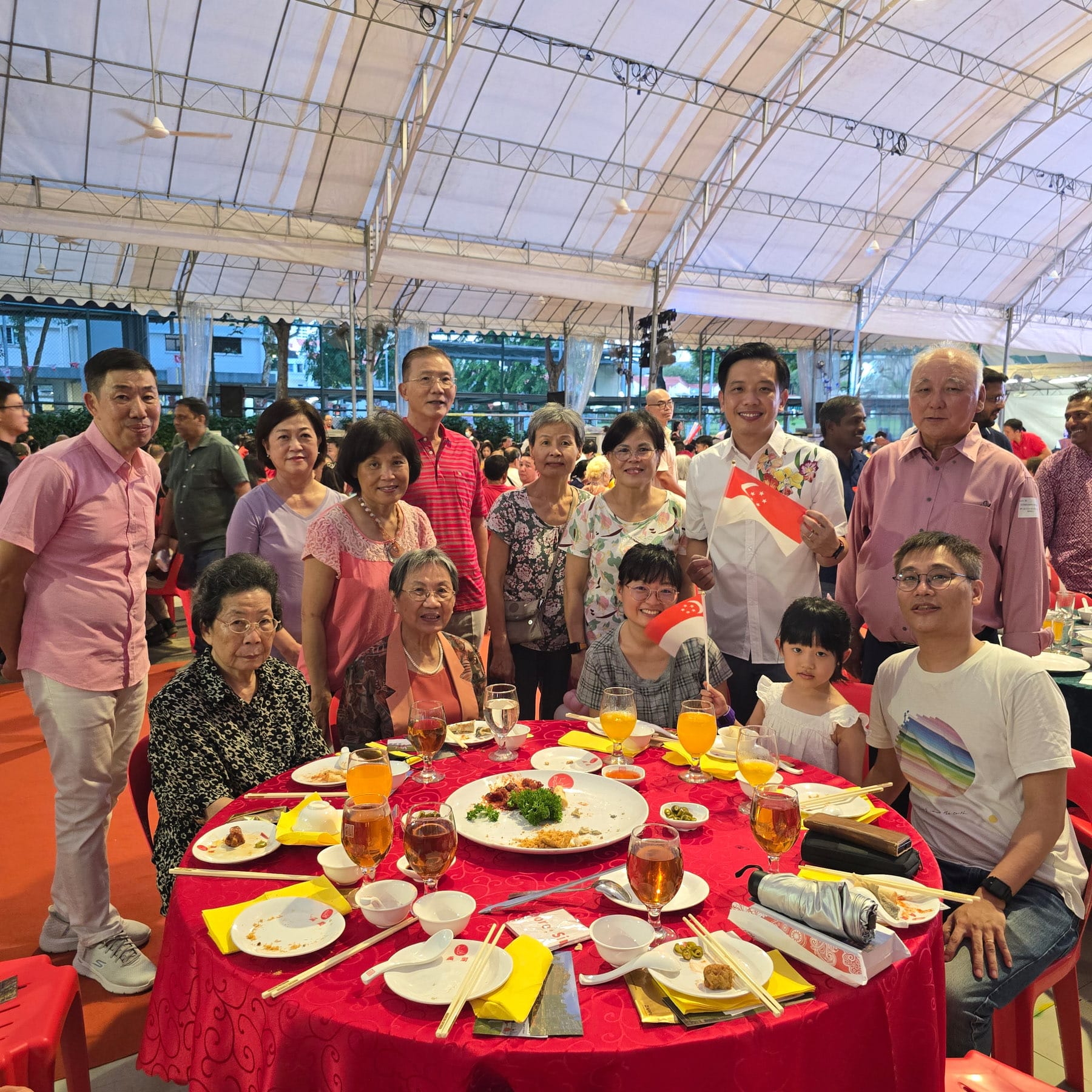 A group of people poses around a red banquet table under a tent, some holding small Singapore flags.