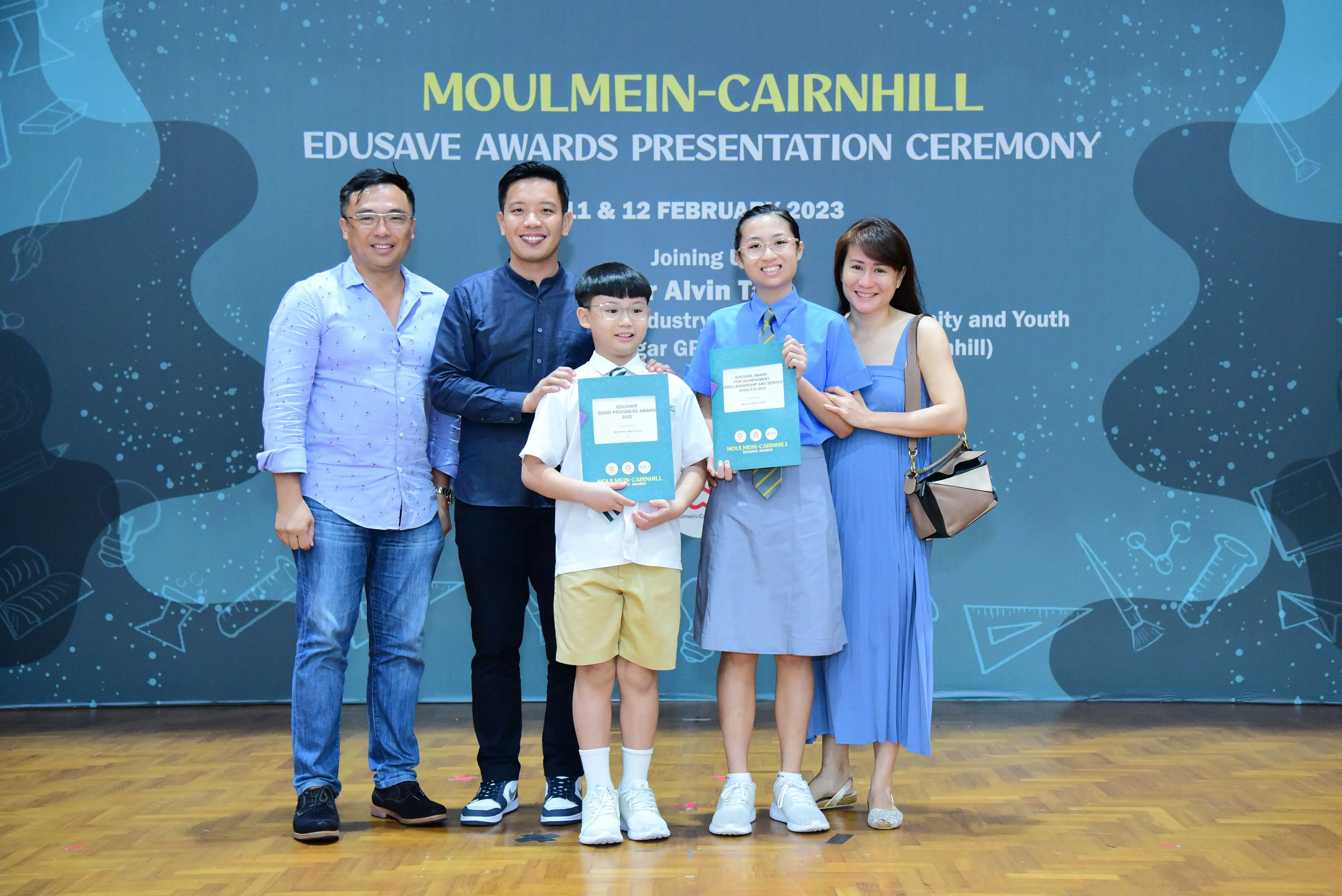 Five people pose at the Moulmein-Cairnhill Edusave Awards Ceremony, two children holding teal award certificates.
