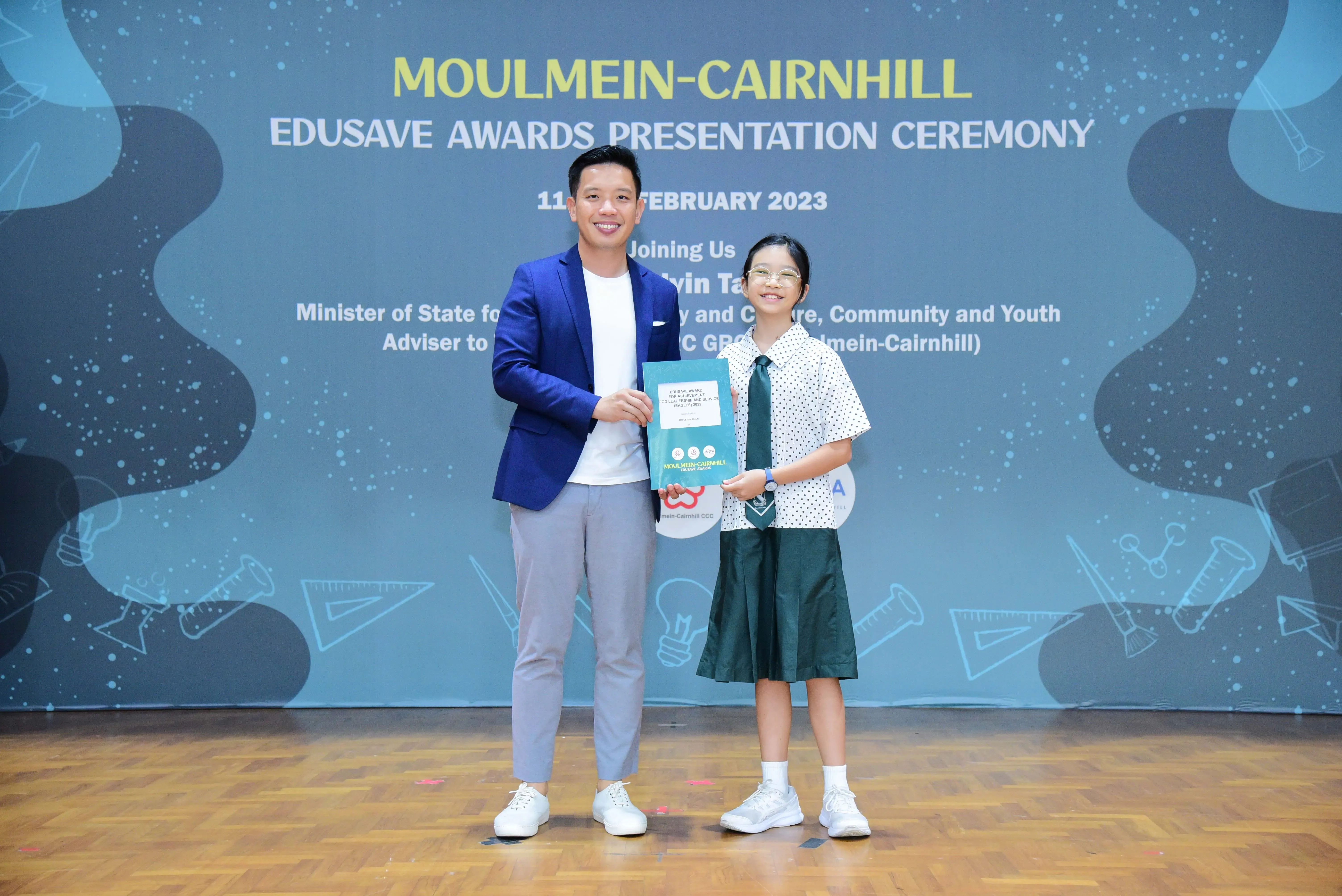 A man in a blue blazer and a young girl in school uniform hold a certificate at the Moulmein-Cairnhill Edusave Awards 2023.