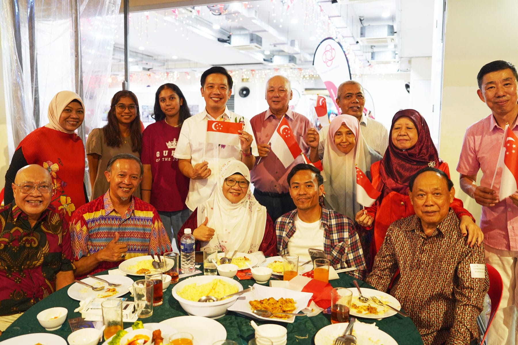 A group of smiling people pose at a dining table, several holding small Singapore flags, with food and drinks visible.