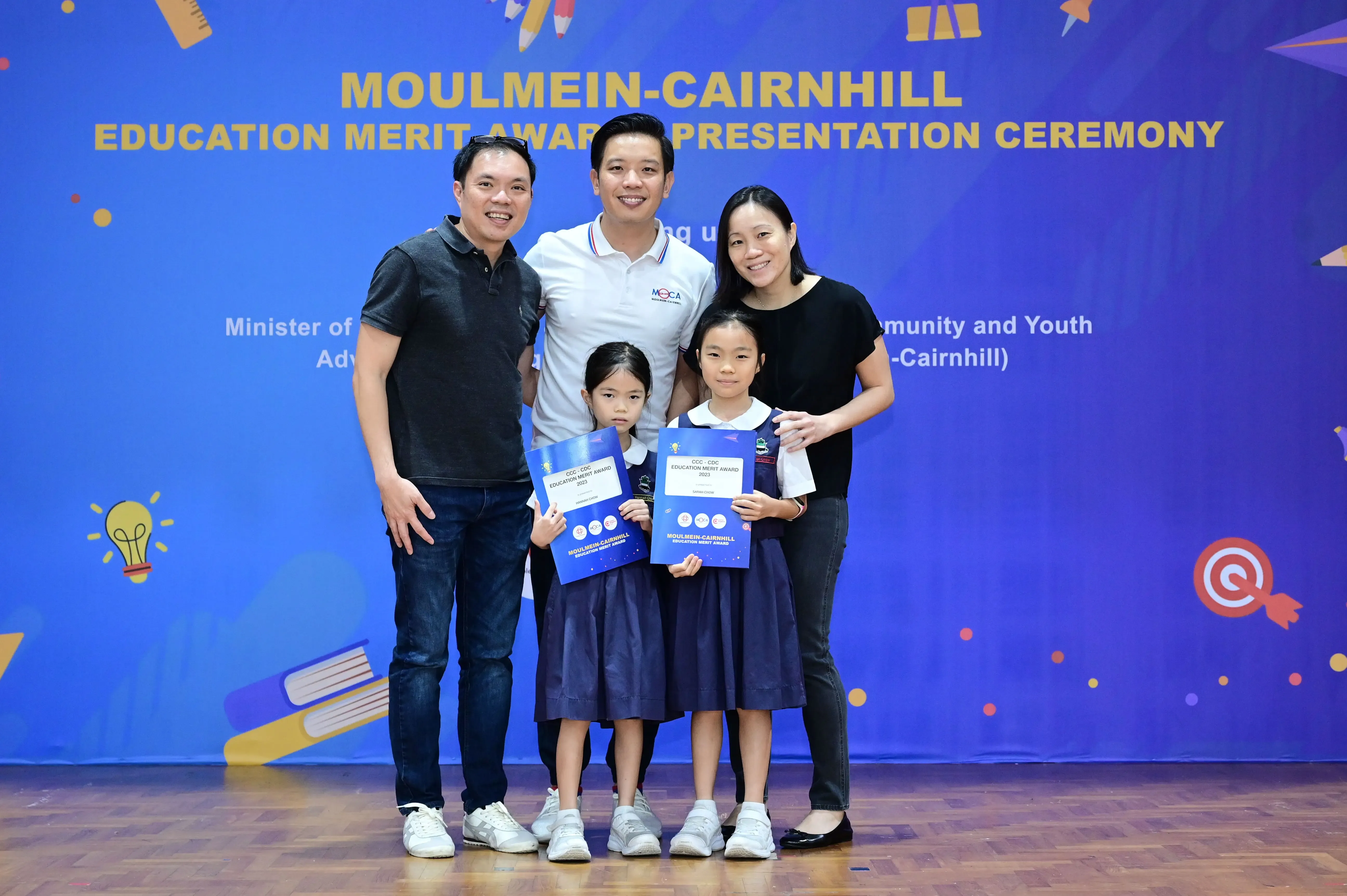 Three adults and two girls in school uniforms holding awards at the Moulmein-Cairnhill Education Merit Award Ceremony.