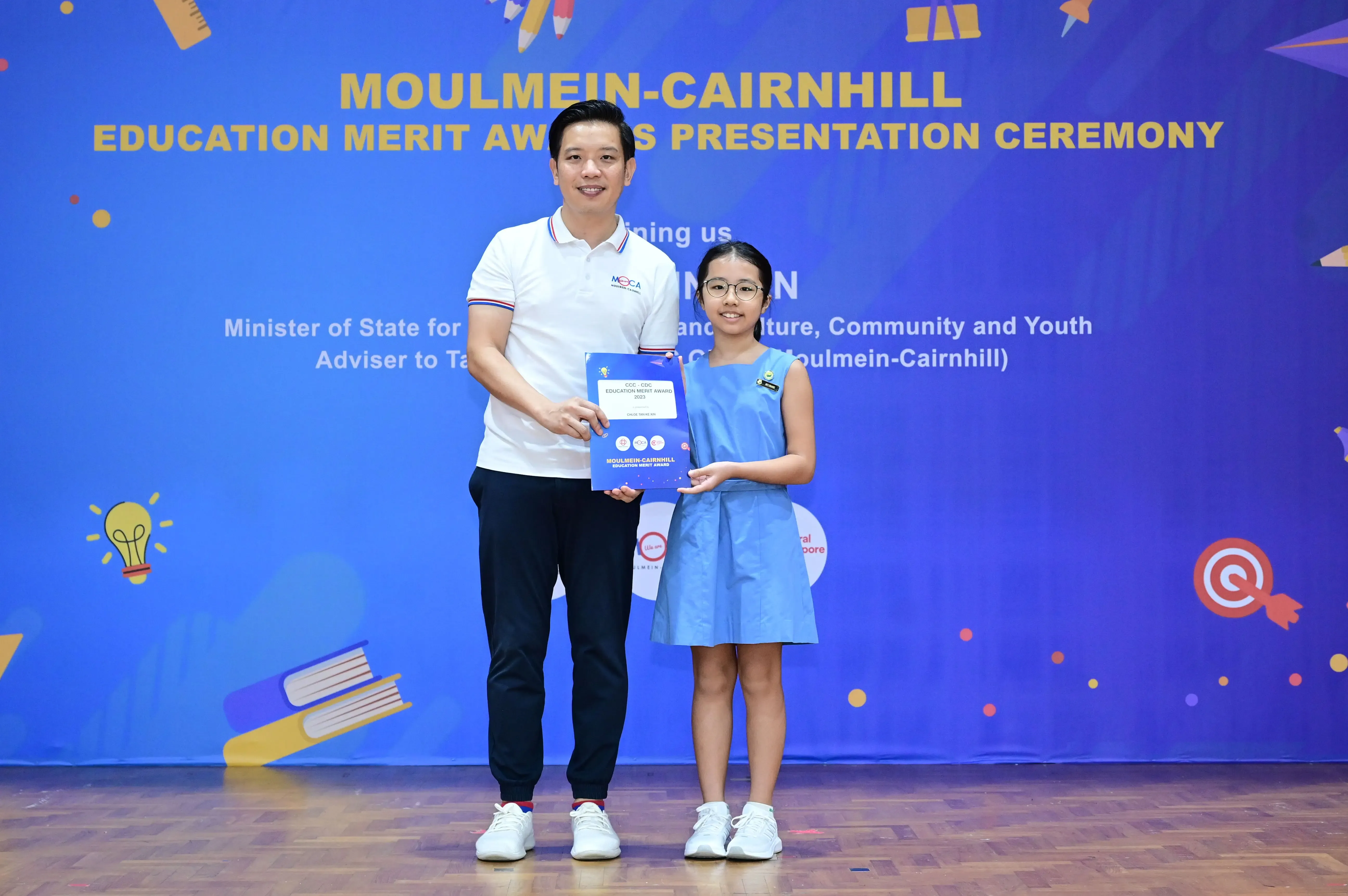 A man and a young girl hold a blue award folder at the Moulmein-Cairnhill Education Merit Award Presentation Ceremony.