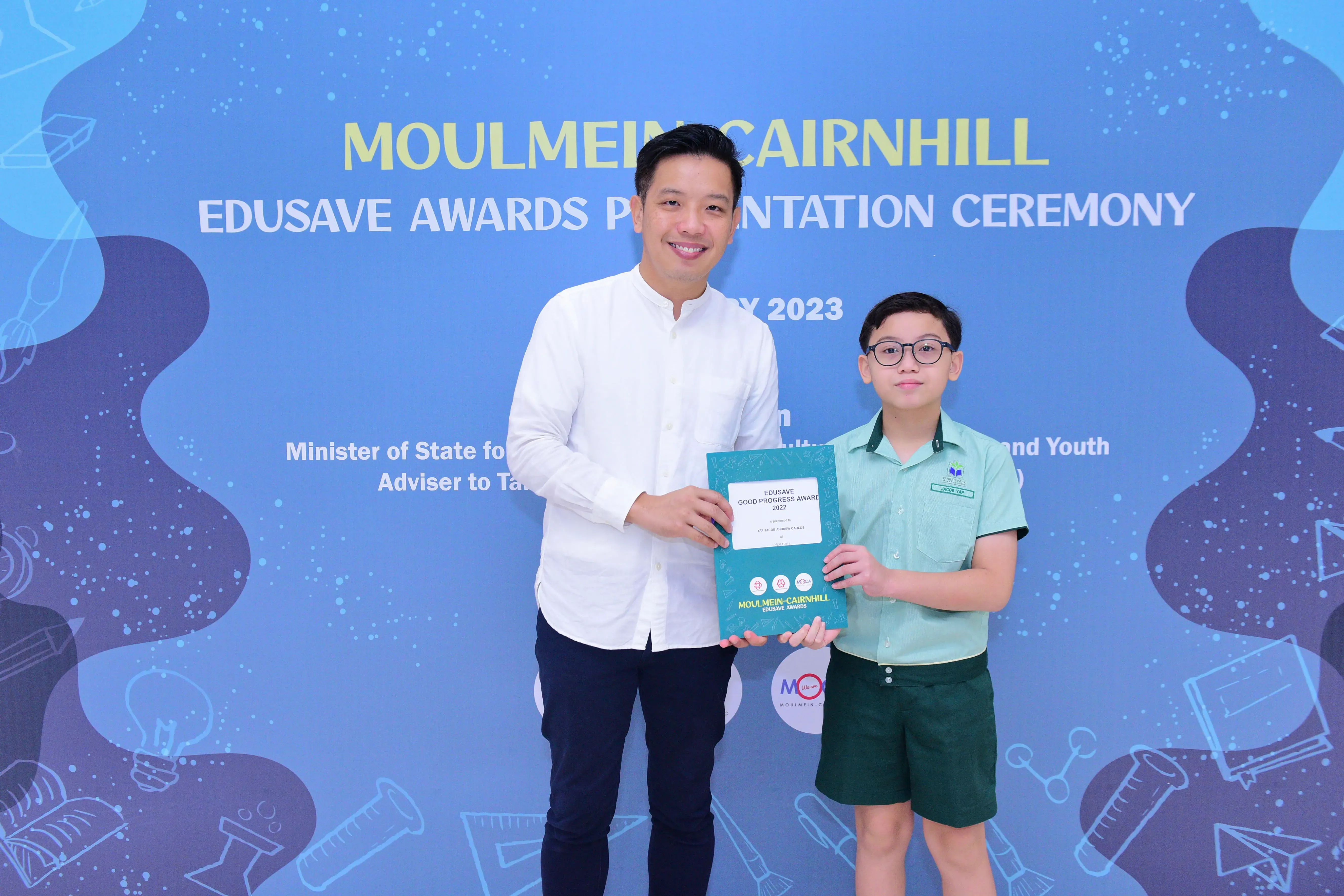 A man and a young boy hold a teal certificate together at the Moulmein-Cairnhill Edusave Awards Ceremony 2023.